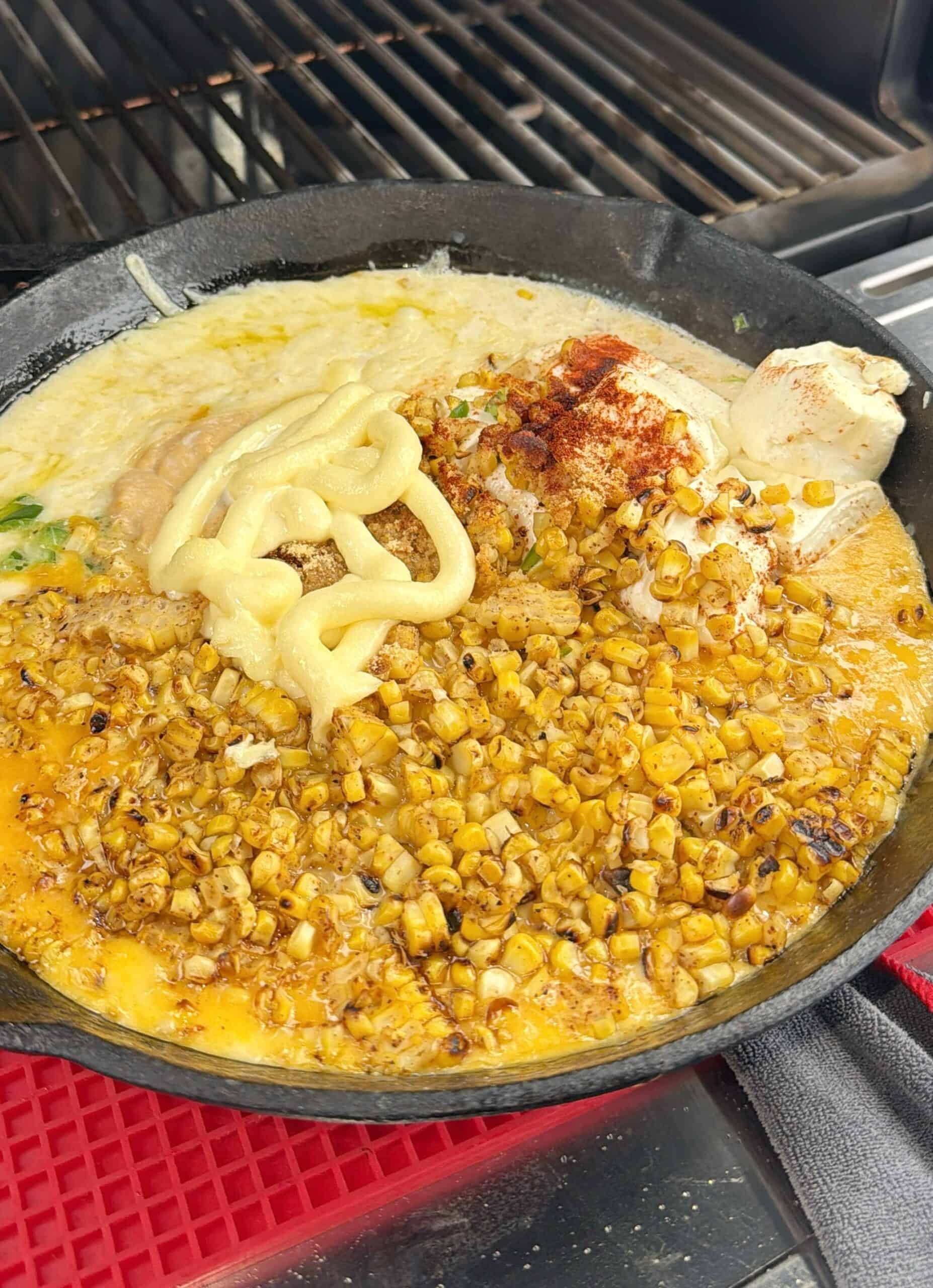 A cast iron skillet on a grill contains charred corn, shredded cheese, mayonnaise, cream cheese, spices, and other ingredients, partially mixed, likely being prepared as a dip.