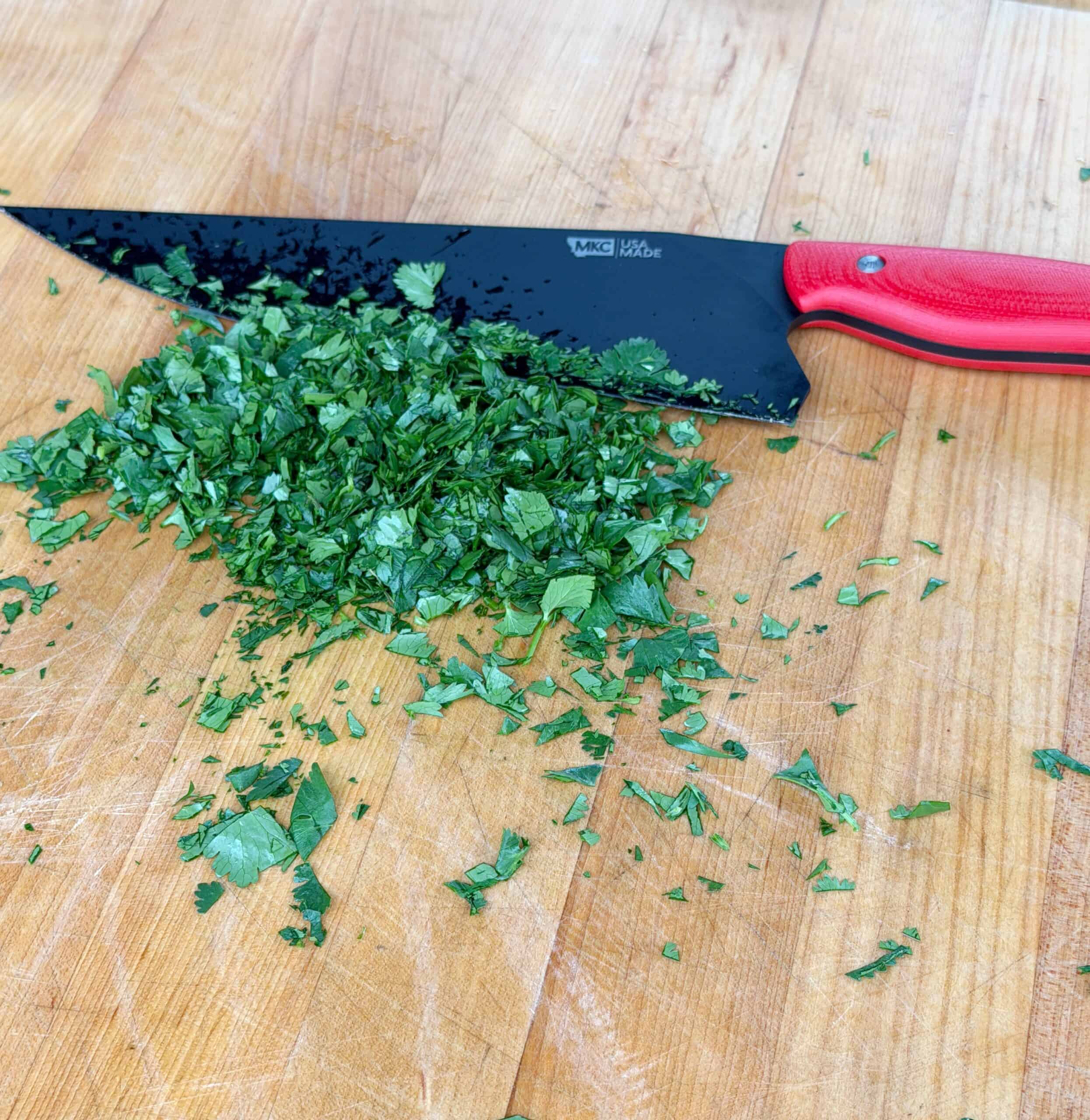 A pile of freshly chopped herbs sits on a wooden cutting board next to a large black kitchen knife with a red handle.