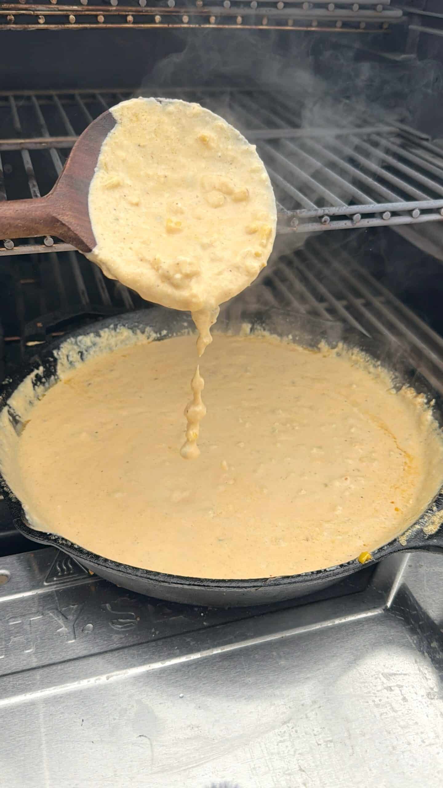 A wooden spoon lifts up a thick, creamy yellow mixture from a cast iron skillet, with some of the mixture dripping back into the pan. The skillet sits on a grill with metal grates above it.