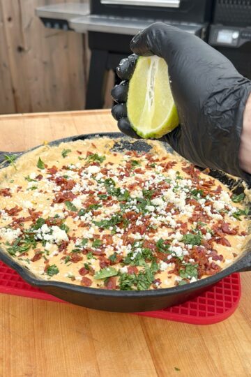 A hand in a black glove squeezes a lime over a skillet filled with a cheesy, bacon and herb-topped dip on a wooden table. A grill is visible in the background.