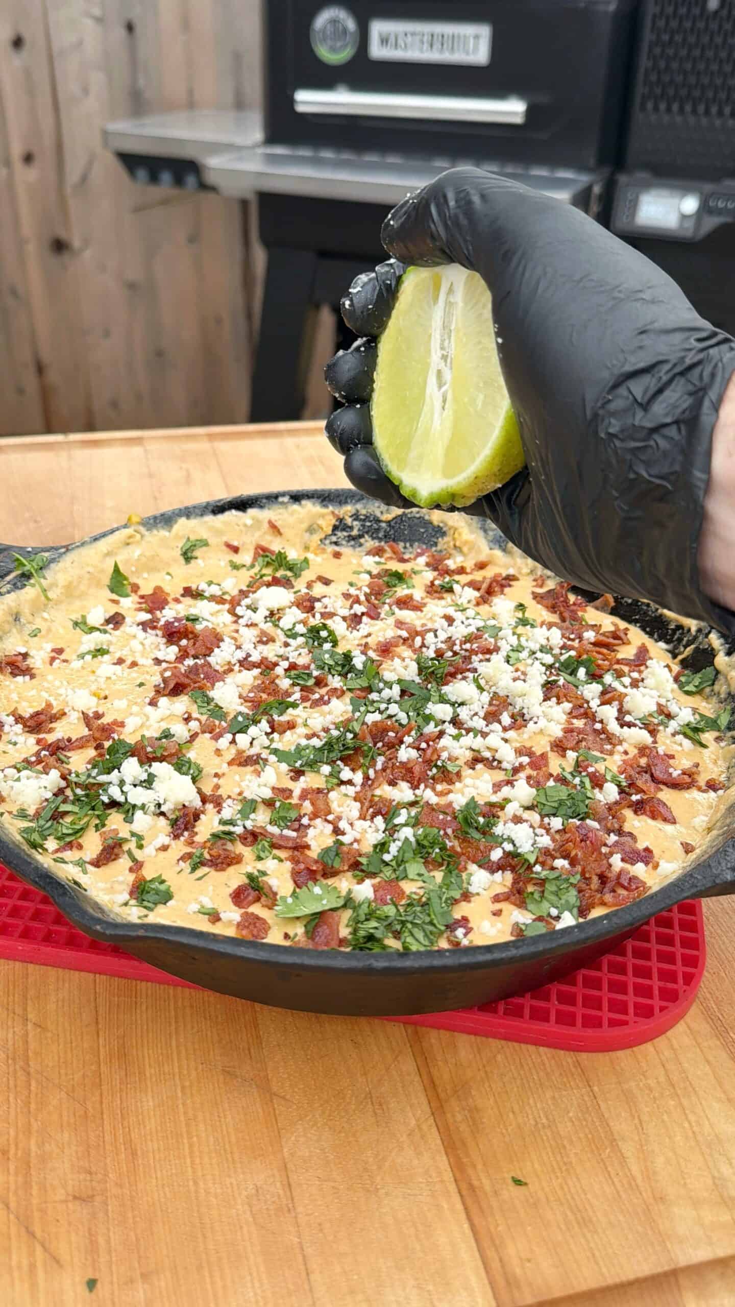 A hand in a black glove squeezes a lime over a skillet filled with a cheesy, bacon and herb-topped dip on a wooden table. A grill is visible in the background. Recipe is for smoked elote dip