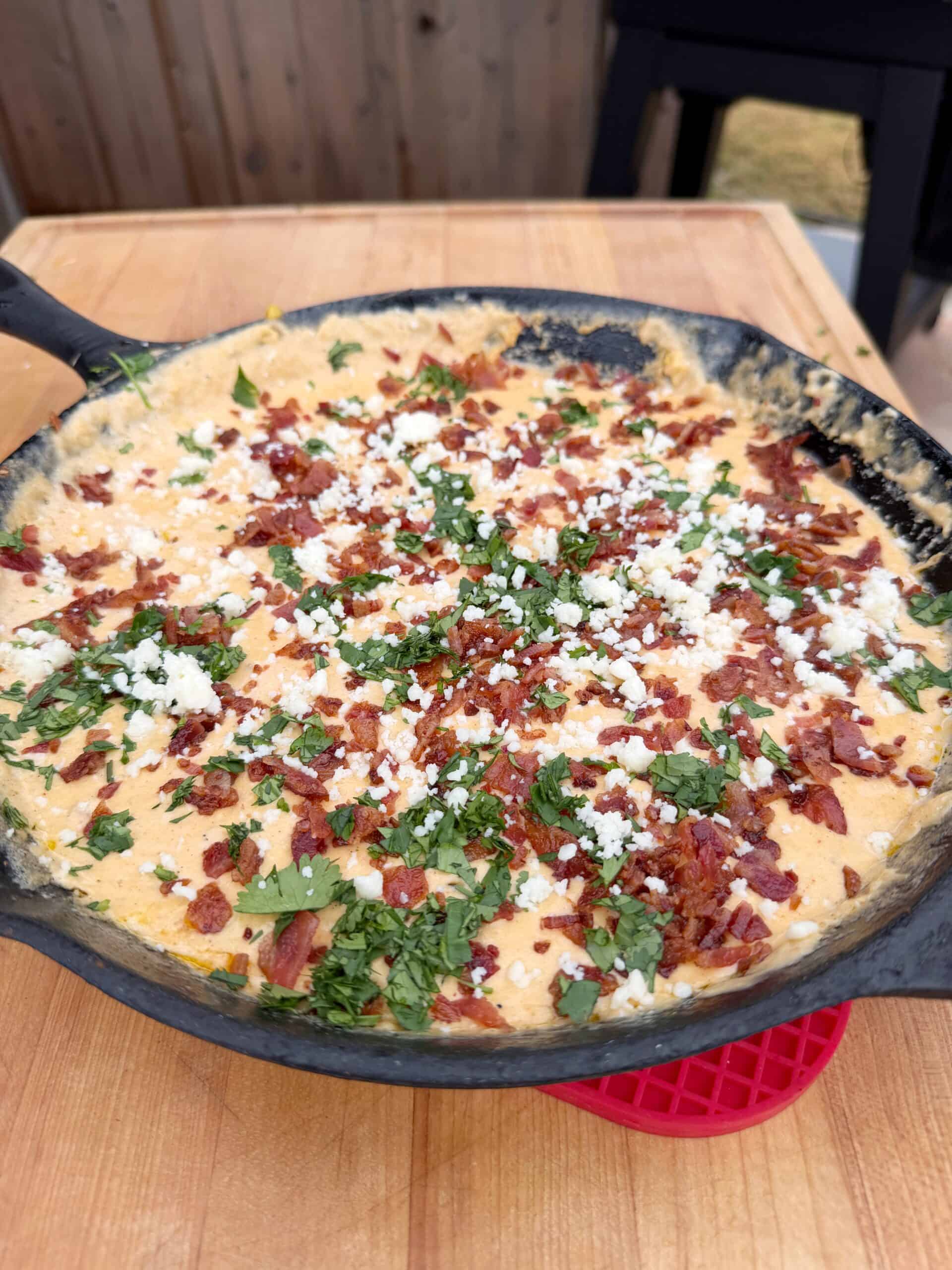 A cast iron skillet filled with creamy dip topped with crumbled bacon, fresh cilantro, and white cheese, sitting on a wooden table.