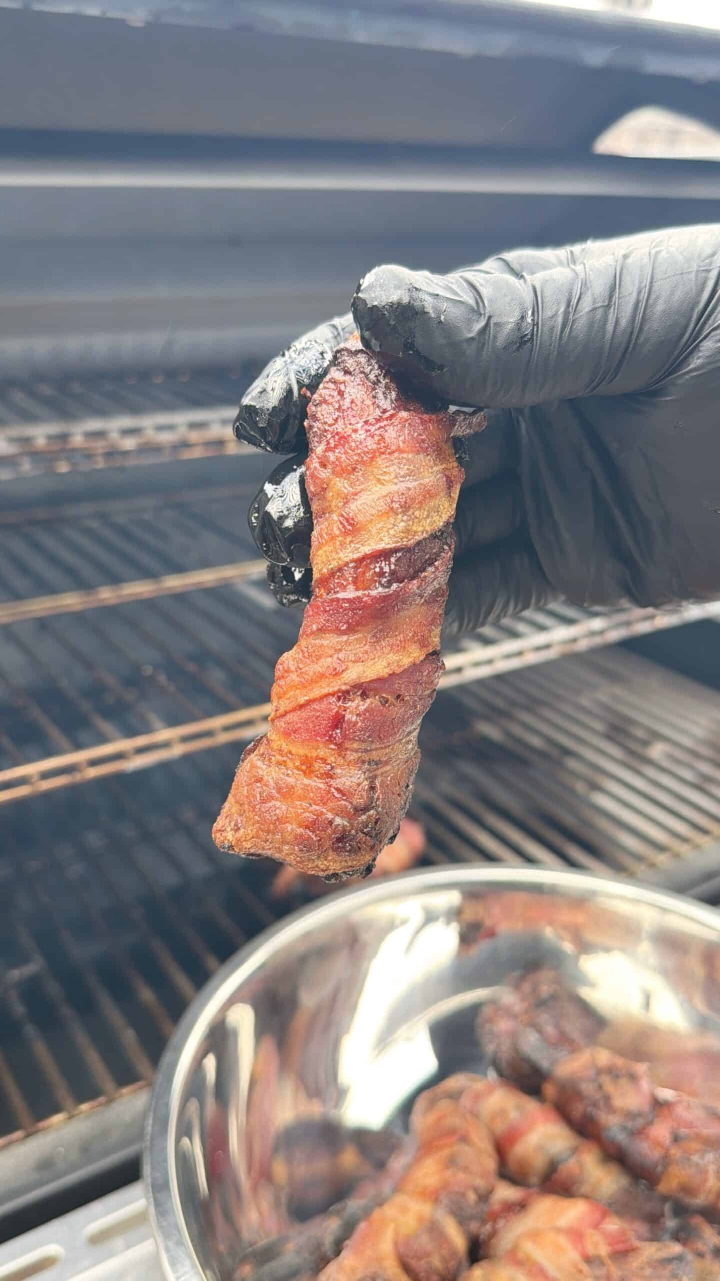A gloved hand holds a piece of smoked, bacon-wrapped meat in front of an open grill, with more pieces in a metal bowl below.