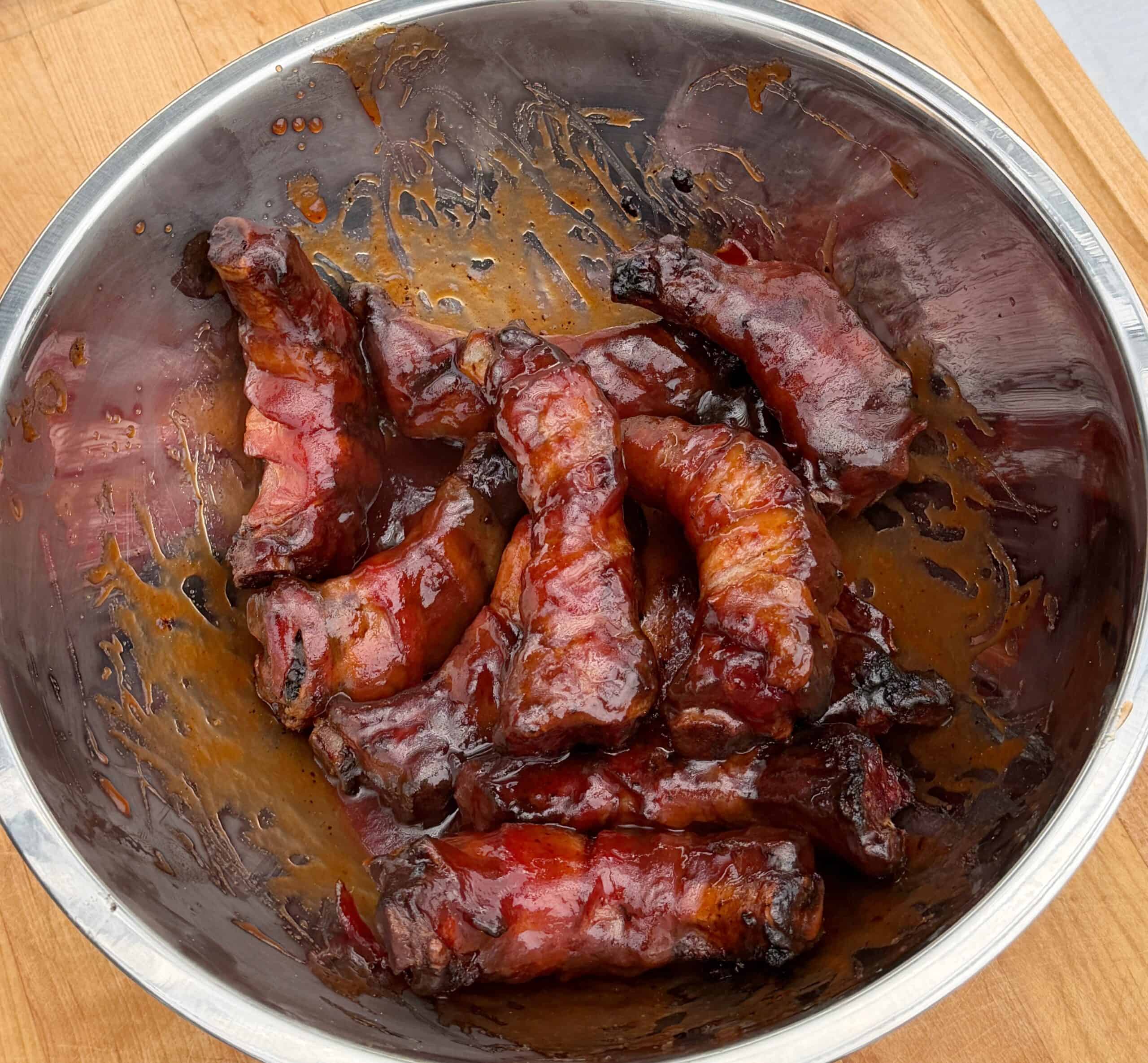 A stainless steel bowl containing several pieces of glazed, cooked bacon wrapped party ribs with a shiny, caramelized coating, resting on a wooden surface.