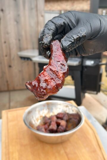A gloved hand holds up a piece of glazed, smoked meat in front of a bowl filled with more meat pieces. An outdoor grill and wooden wall are blurred in the background.