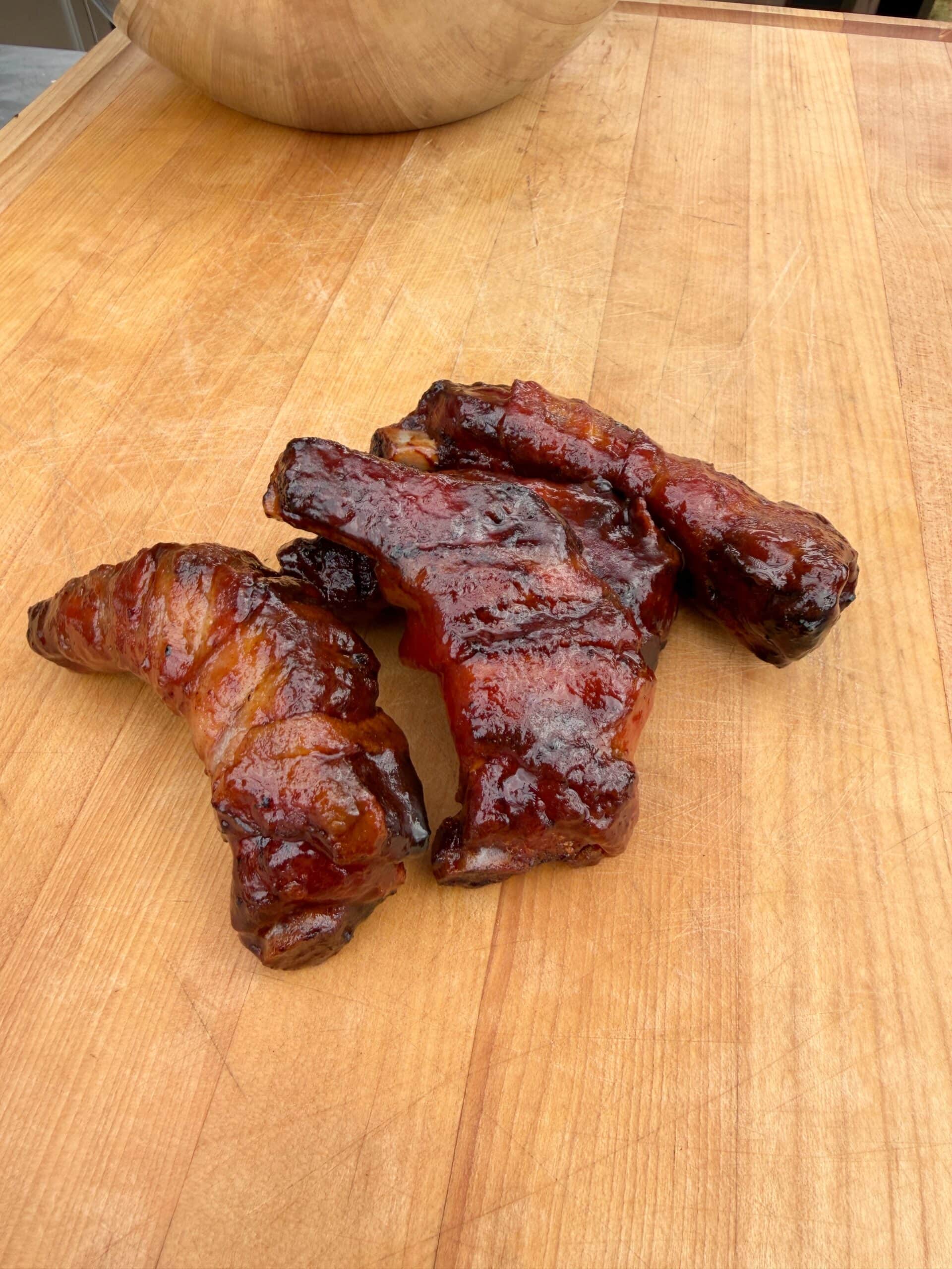 Three pieces of glazed, barbecued pork ribs rest on a wooden cutting board. The meat is caramelized and shiny, showing a rich, reddish-brown color with visible grill marks.