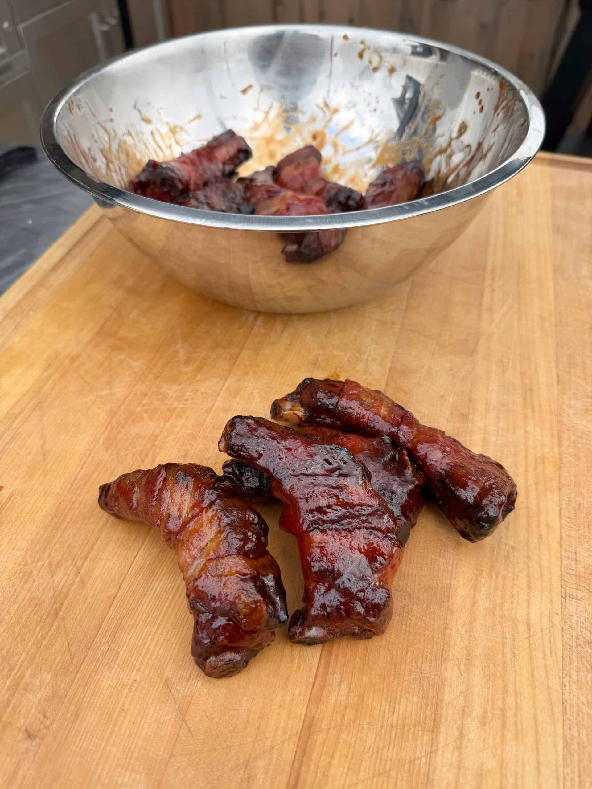 Three pieces of glazed, cooked meat are arranged on a wooden cutting board, with a metal bowl containing more pieces of meat in the background. The meat appears shiny and caramelized.