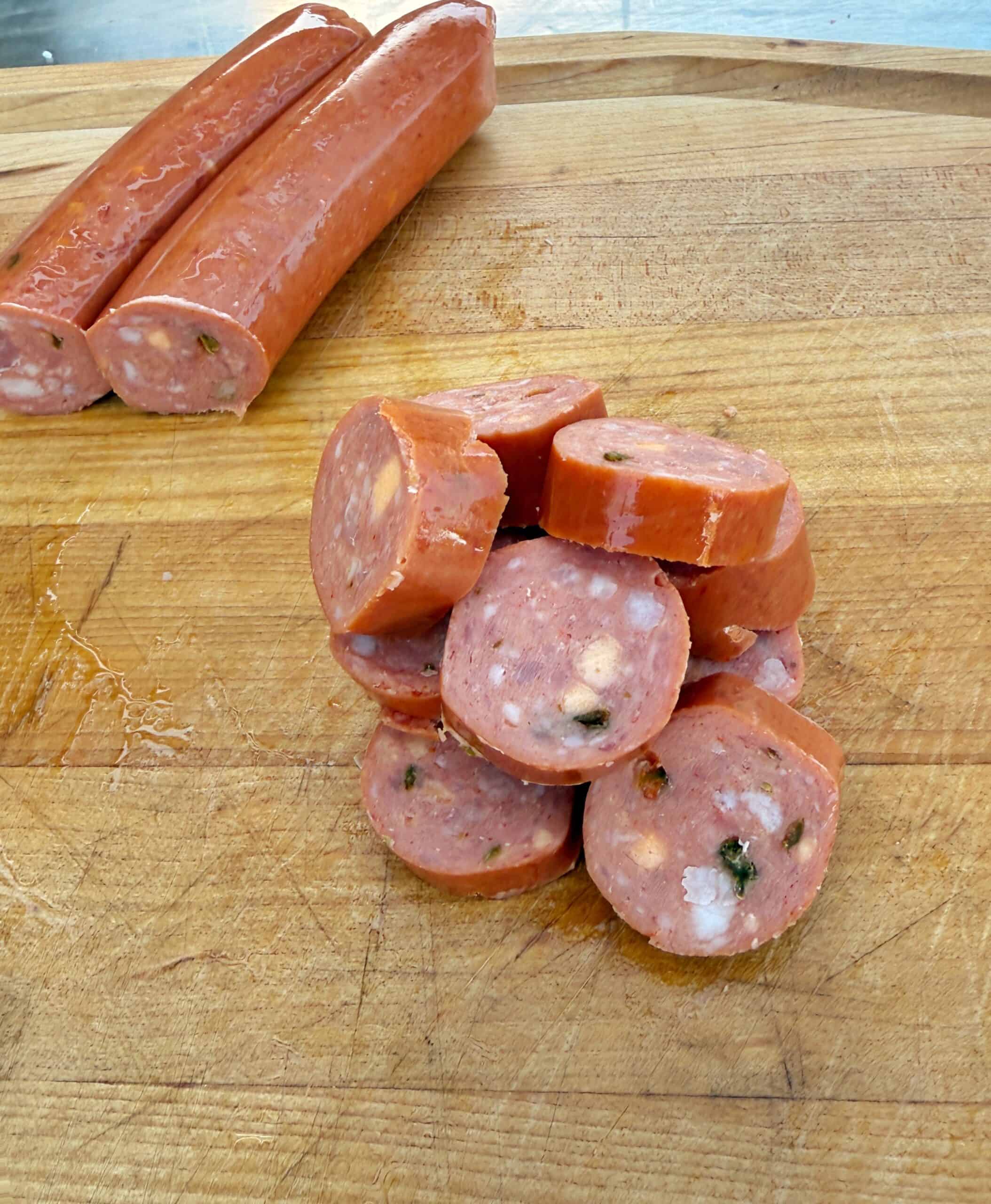 Sliced sausage with visible herbs and cheese on a wooden cutting board, with two unsliced sausage links in the background.