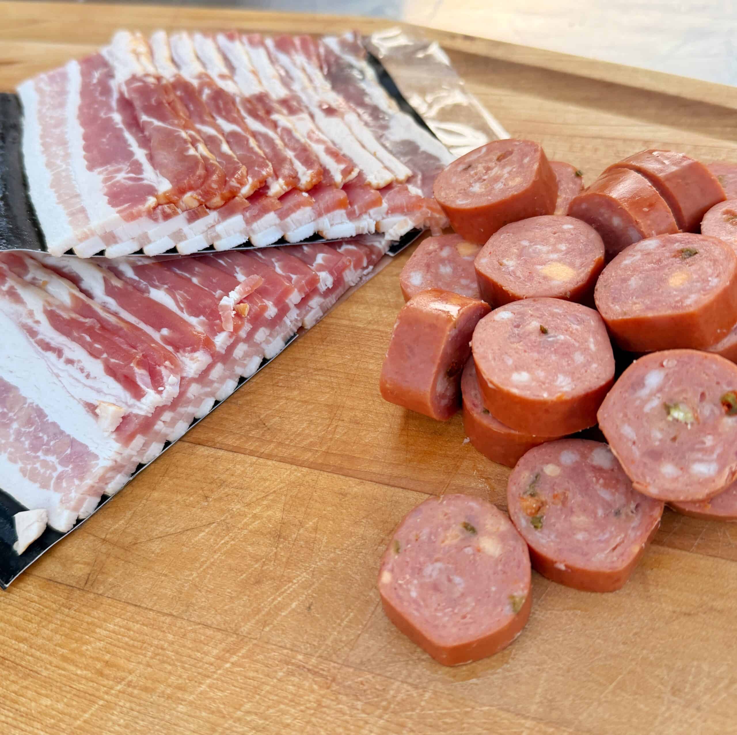 Sliced raw bacon and several round slices of sausage are arranged on a wooden cutting board, ready for cooking or preparation.