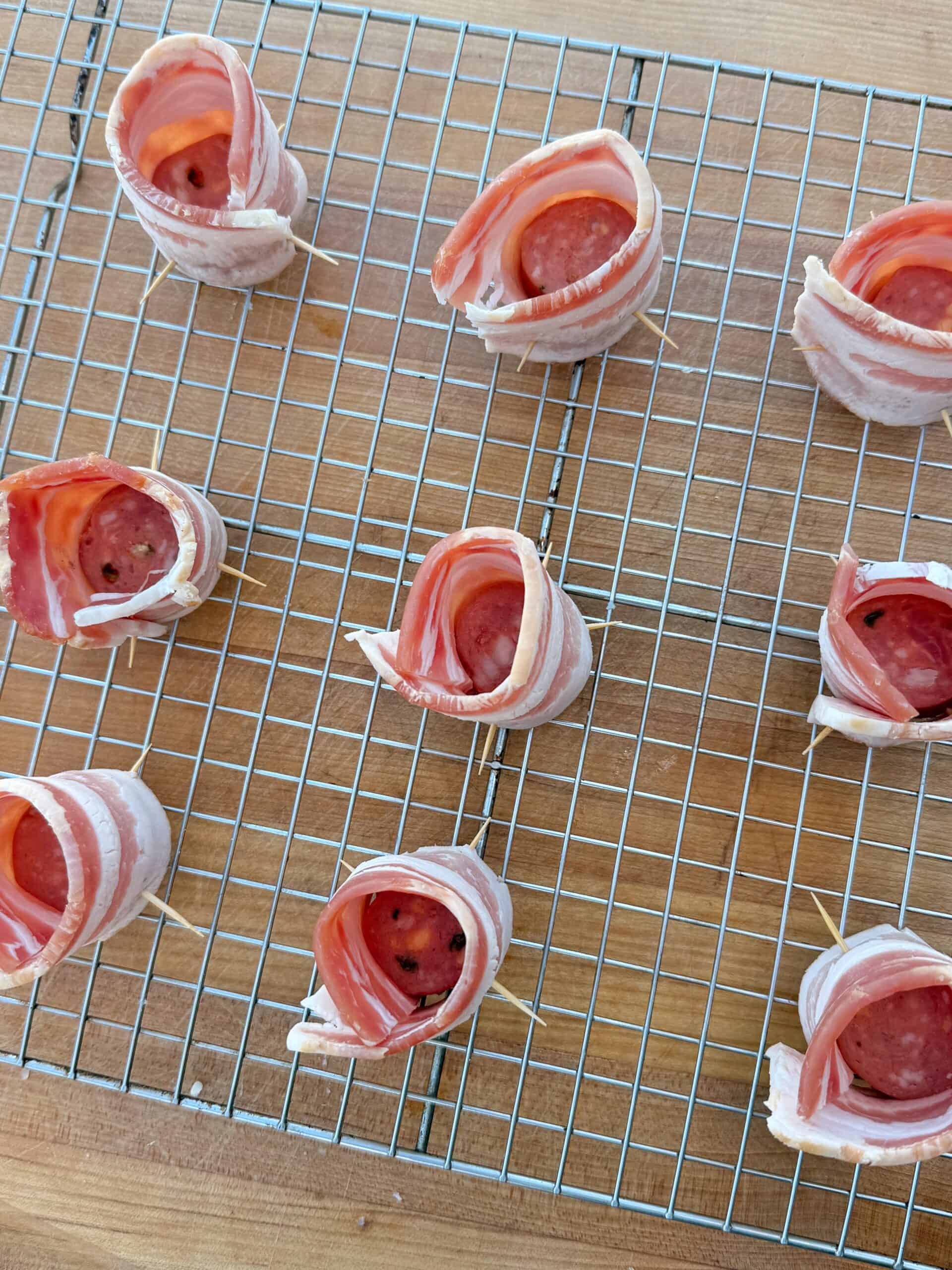 Slices of bacon wrapped around slices of pepperoni and secured with toothpicks are arranged on a metal cooling rack on a wooden surface, ready to be cooked.