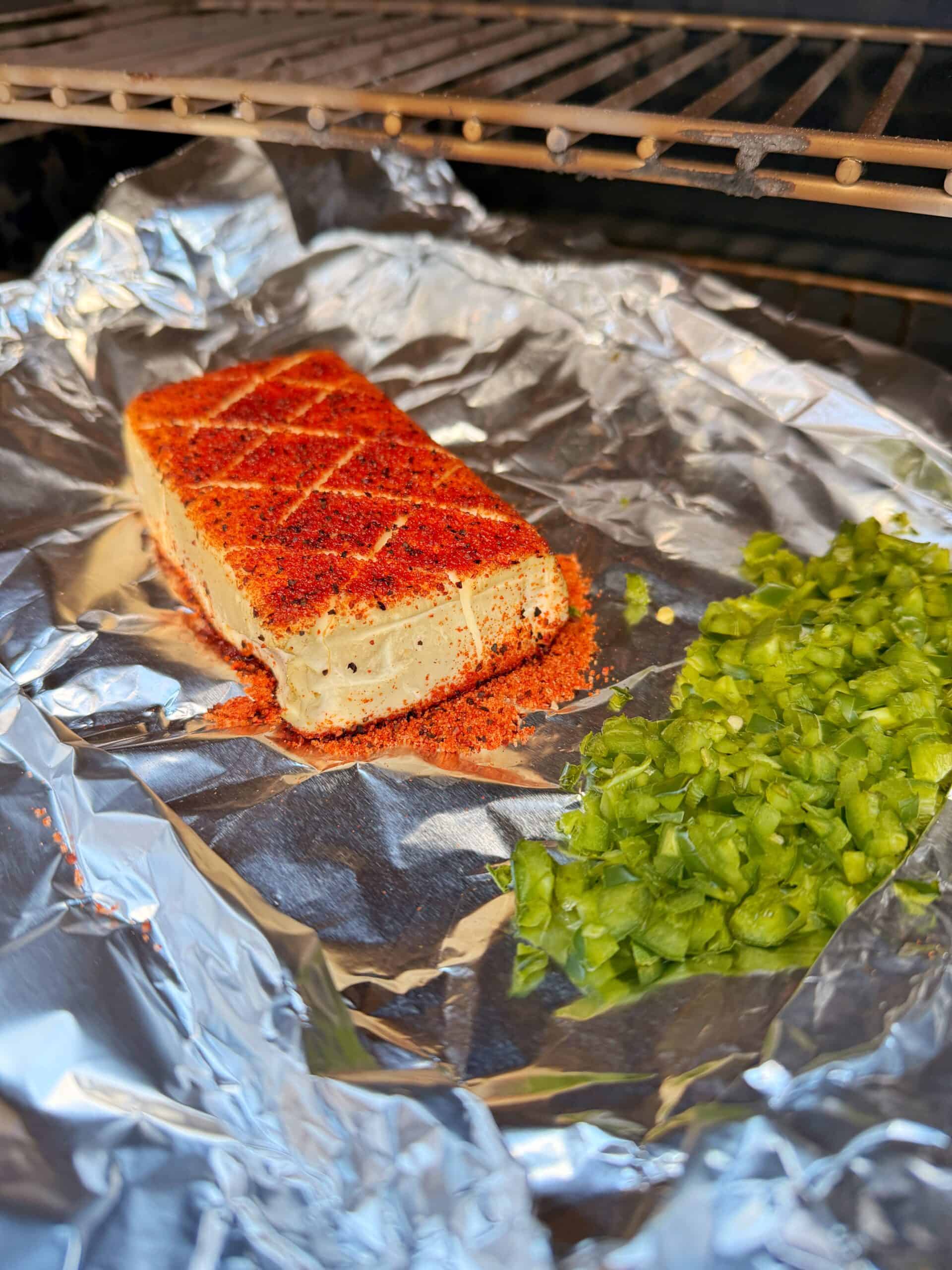 A block of seasoned tofu with a crosshatch pattern sits on aluminum foil next to a pile of chopped green bell peppers, both placed inside an oven.