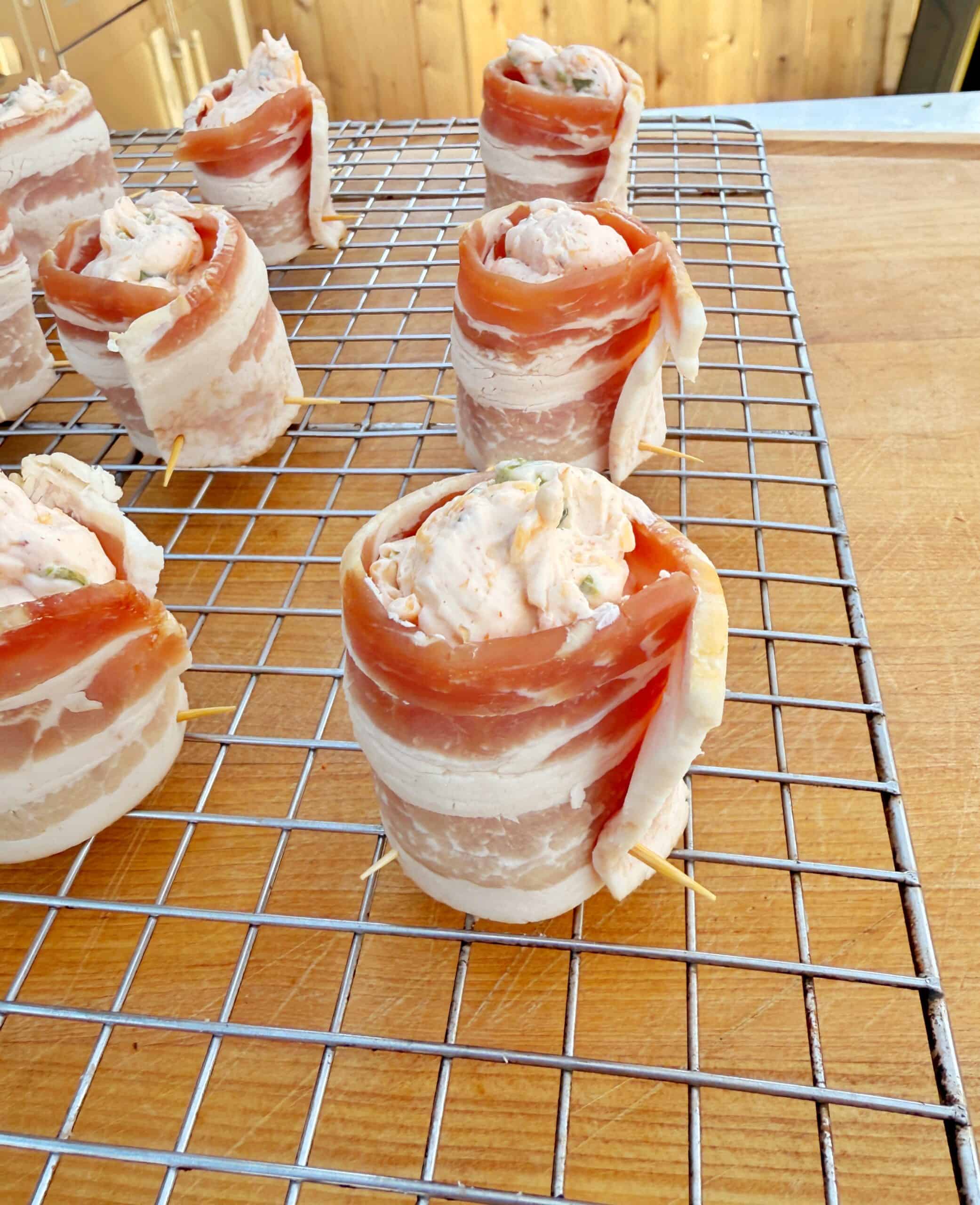 Several jalapeno popper pig shots filled with a creamy cheese mixture are arranged on a wire rack over a wooden surface, ready to be cooked.