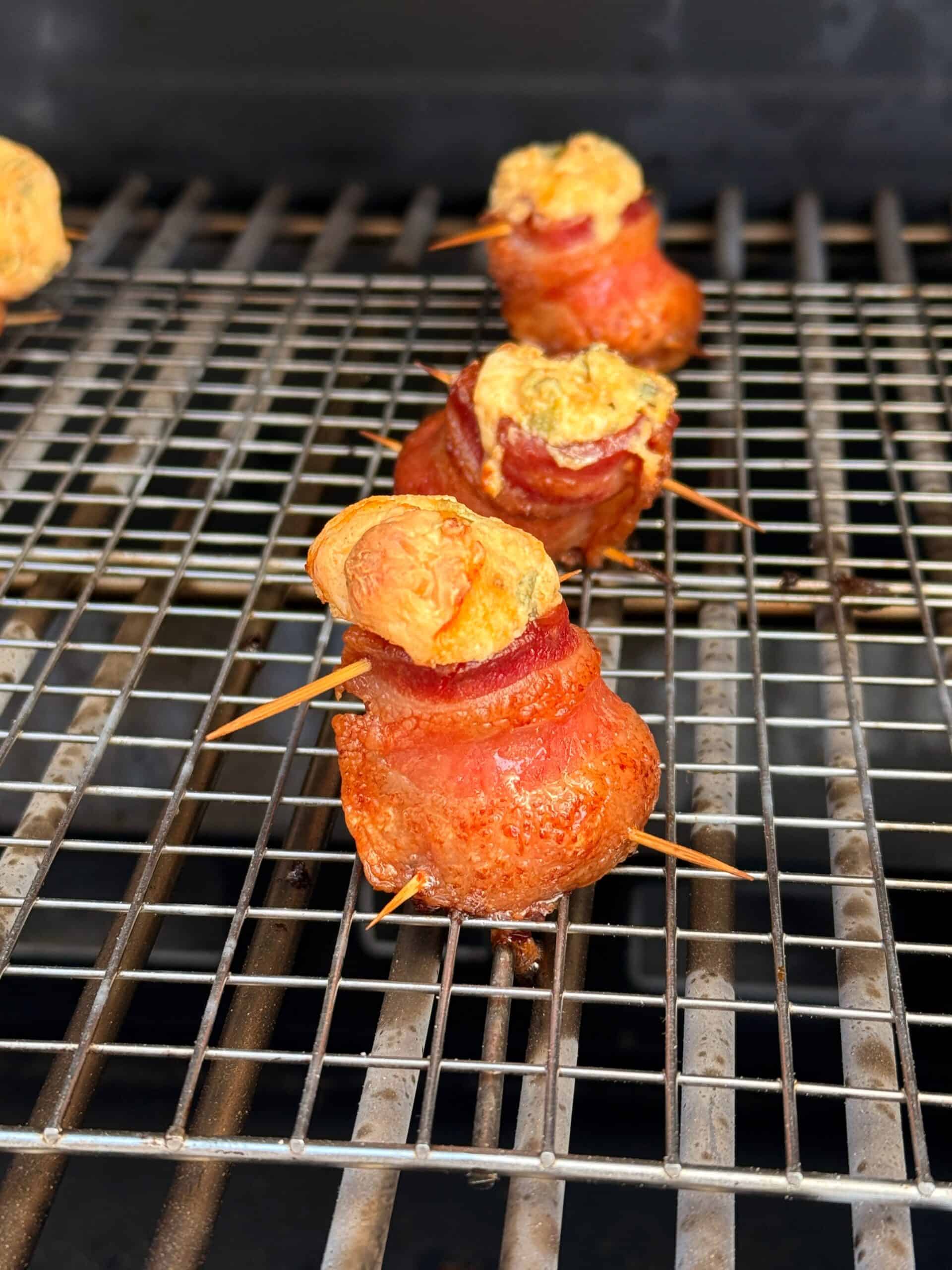Bacon-wrapped jalapeño poppers secured with toothpicks and topped with a golden cheese mixture, arranged on a metal grill rack for cooking.
