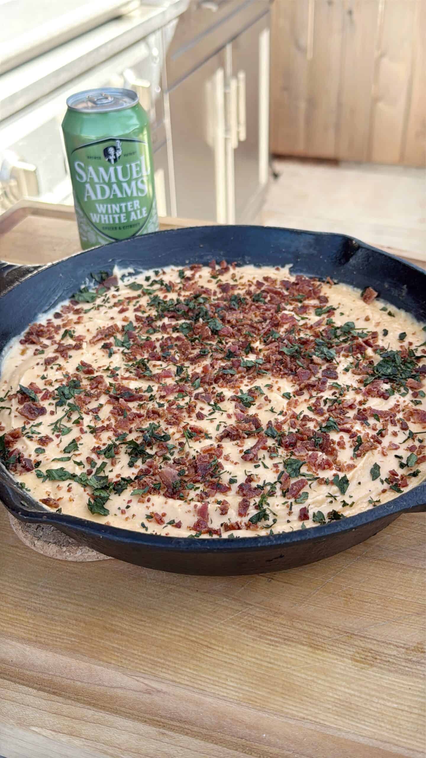 A skillet filled with creamy smoked beer cheese dip, topped with crumbled bacon and chopped herbs, sits on a wooden surface. A can of Samuel Adams Winter Lager is placed in the background.