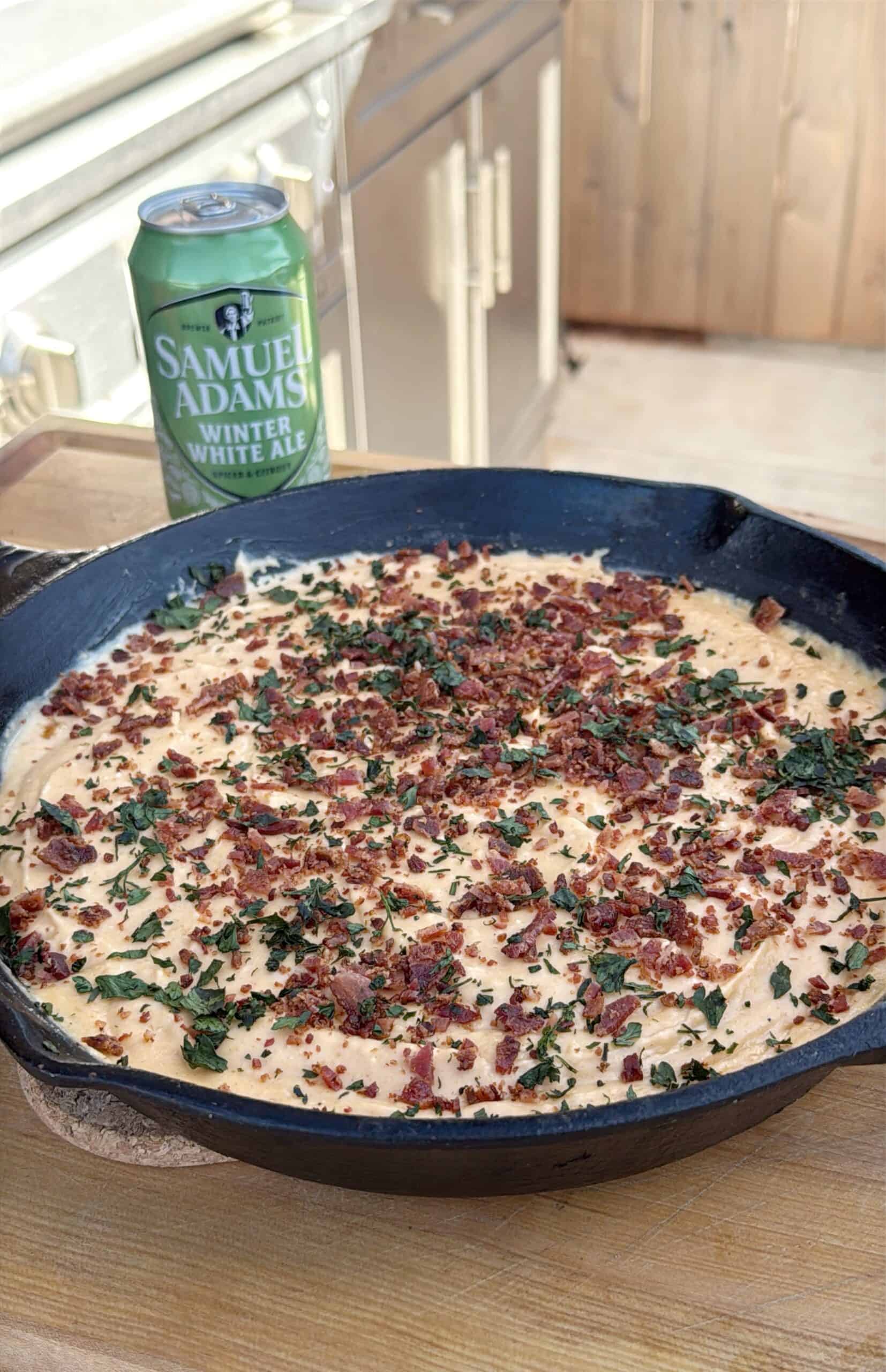 A cast-iron skillet filled with a creamy smoked beer cheese dip topped with chopped bacon and herbs sits on a wooden surface. A can of Samuel Adams Winter White Ale is placed next to the skillet.