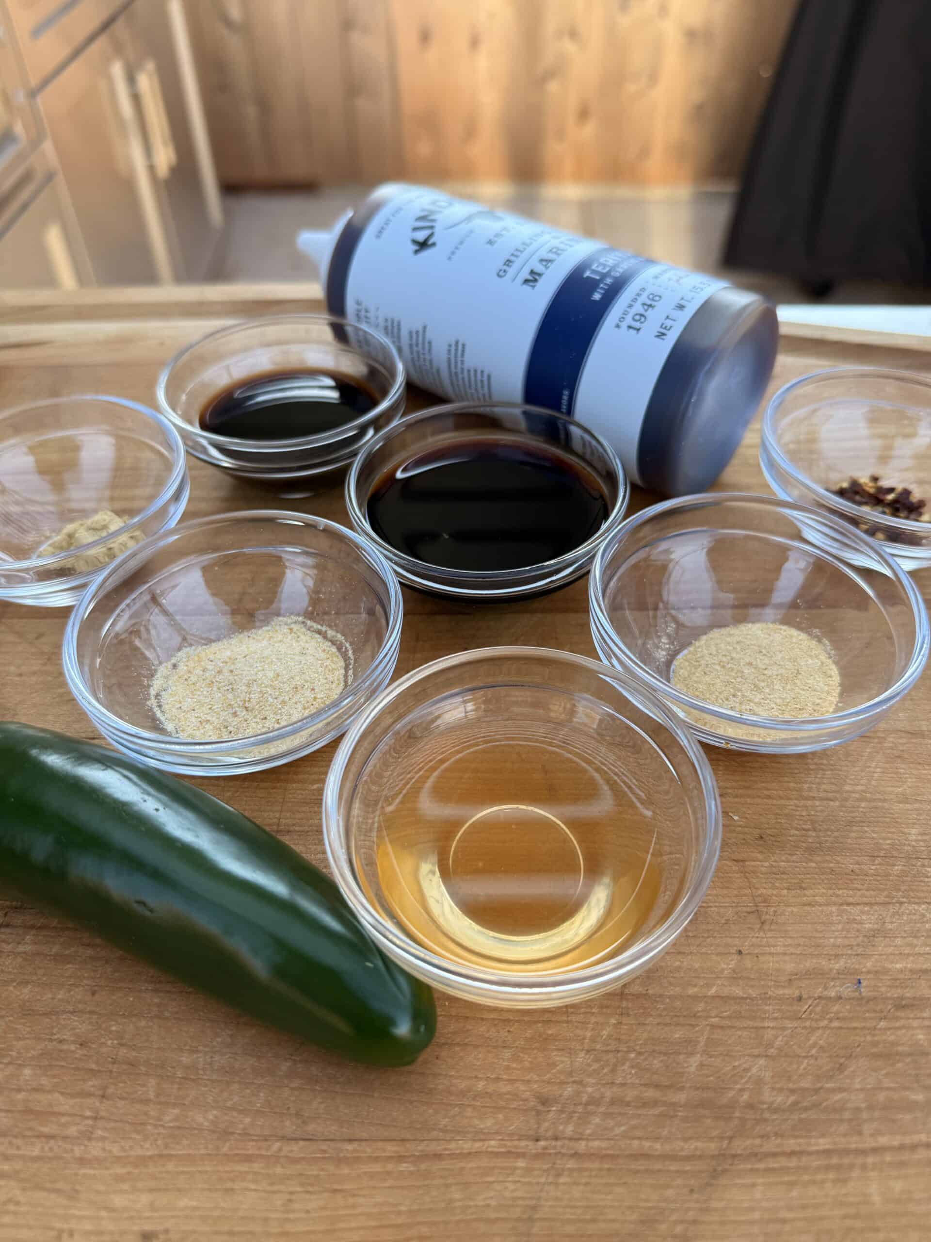 A wooden cutting board holds a fresh jalapeño, small glass bowls filled with spices, soy sauce, vinegar, and a bottle of Kirkland maple syrup lying on its side in the background.
