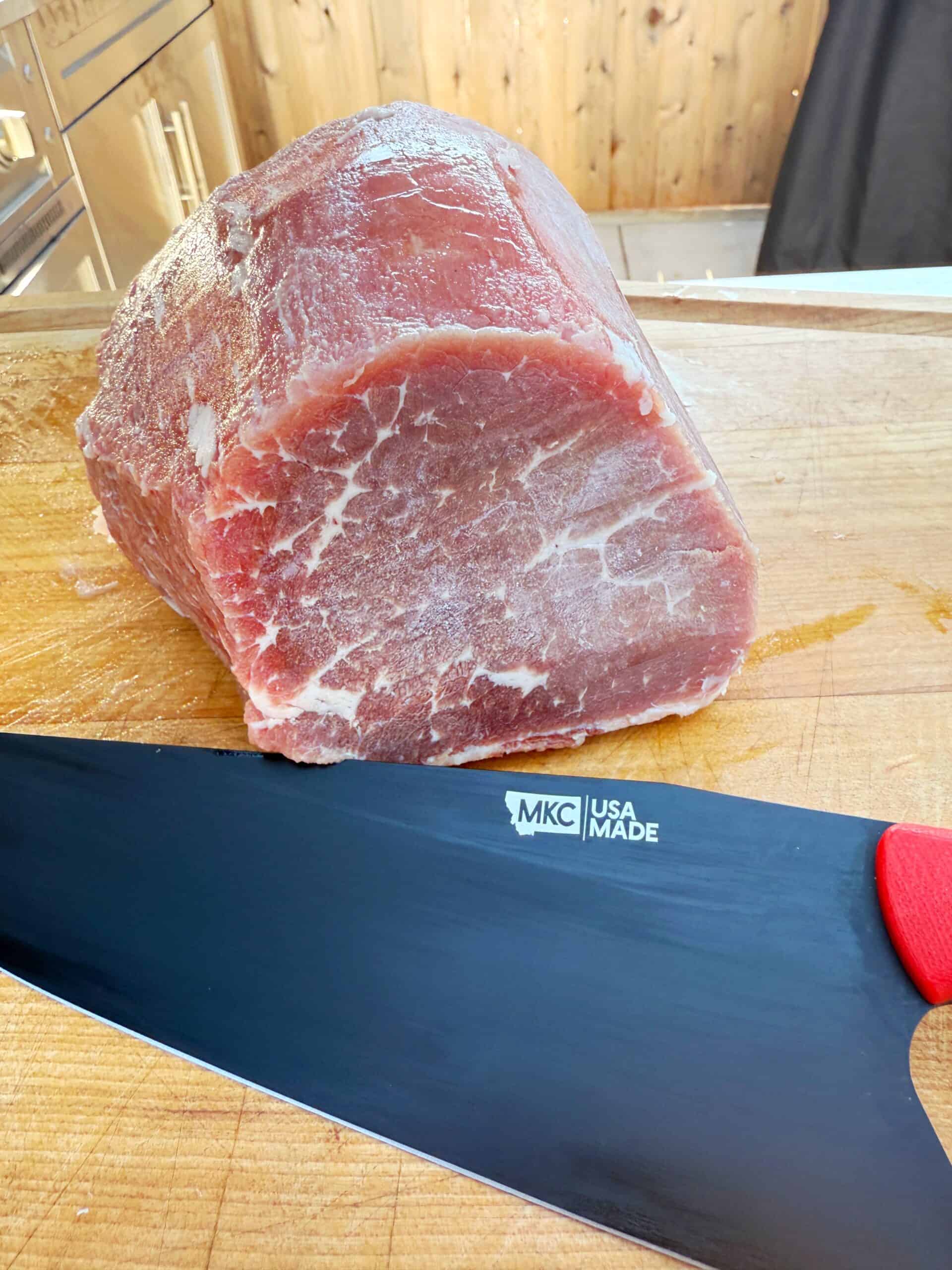 A large piece of raw beef sits on a wooden cutting board next to a black knife with a red handle. The knife blade is labeled “MKC USA MADE.” Wooden cabinets are visible in the background.