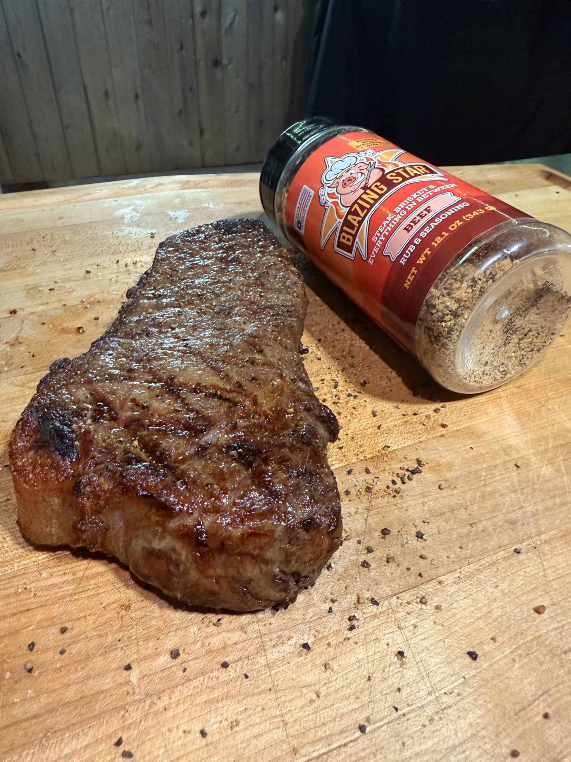 A grilled steak rests on a wooden cutting board next to a tipped-over container of Blazing Star Beef Rub seasoning, with some spice scattered around.