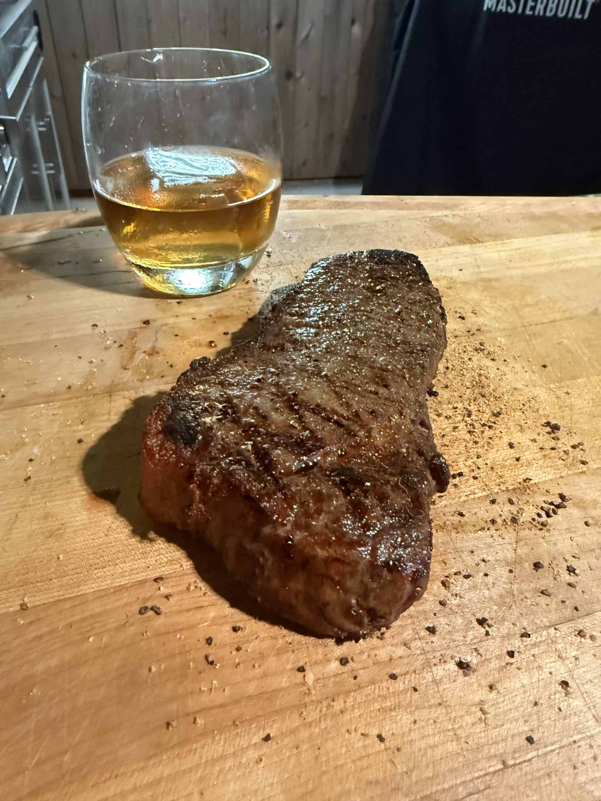 A cooked steak seasoned with pepper rests on a wooden cutting board next to a glass of amber-colored whiskey. A fence and a person wearing a Masterbuilt shirt are in the background.
