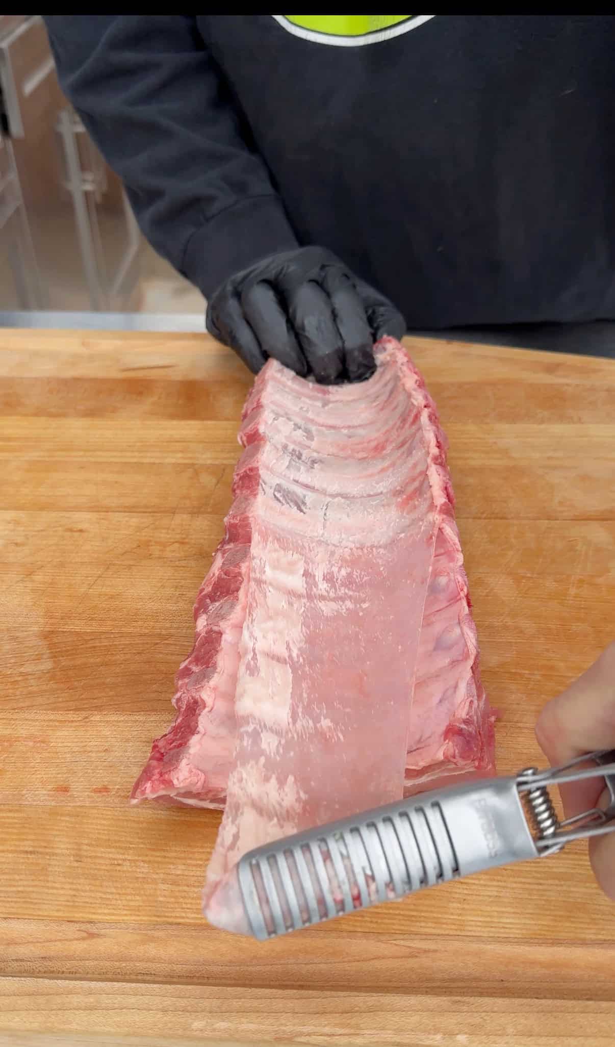 A person wearing black gloves uses a metal tool to remove the thin membrane from a rack of raw pork ribs on a wooden cutting board.