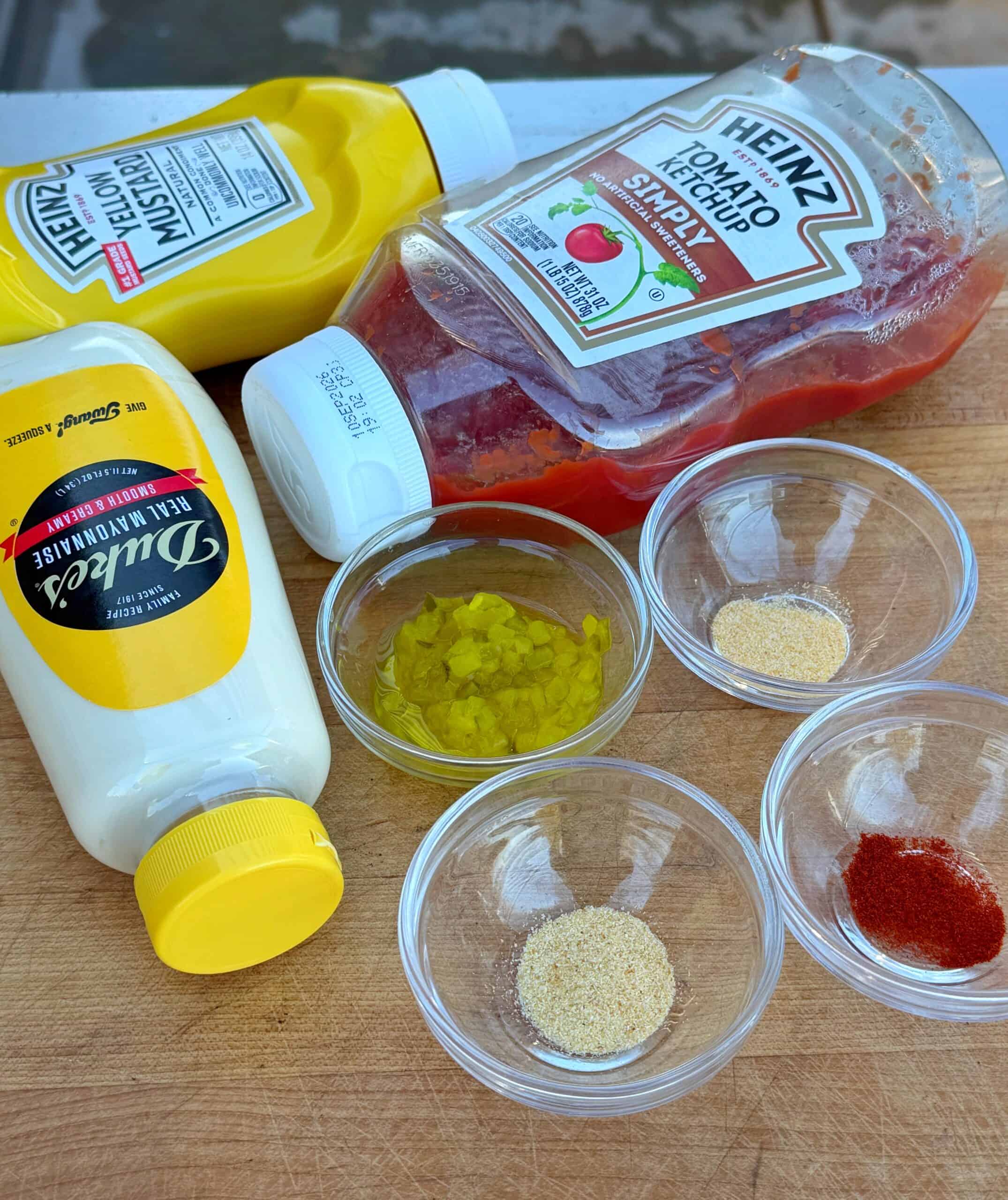 Three condiment bottles (mustard, ketchup, mayonnaise) lie on a wooden surface beside five small bowls containing chopped pickles, garlic powder, onion powder, paprika, and another powdered spice.