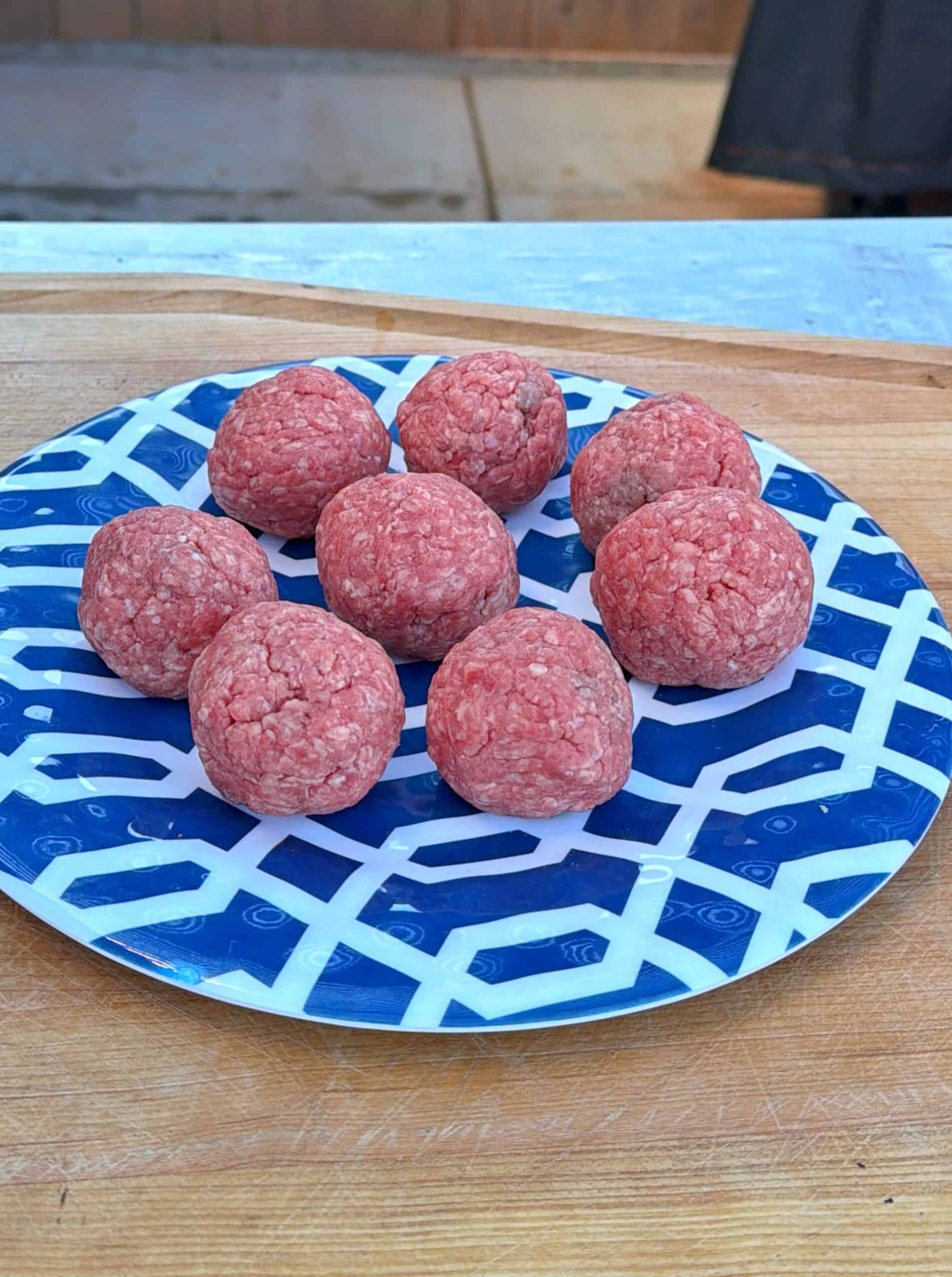 Eight raw ground beef meatballs on a blue and white patterned plate, placed on a wooden surface.