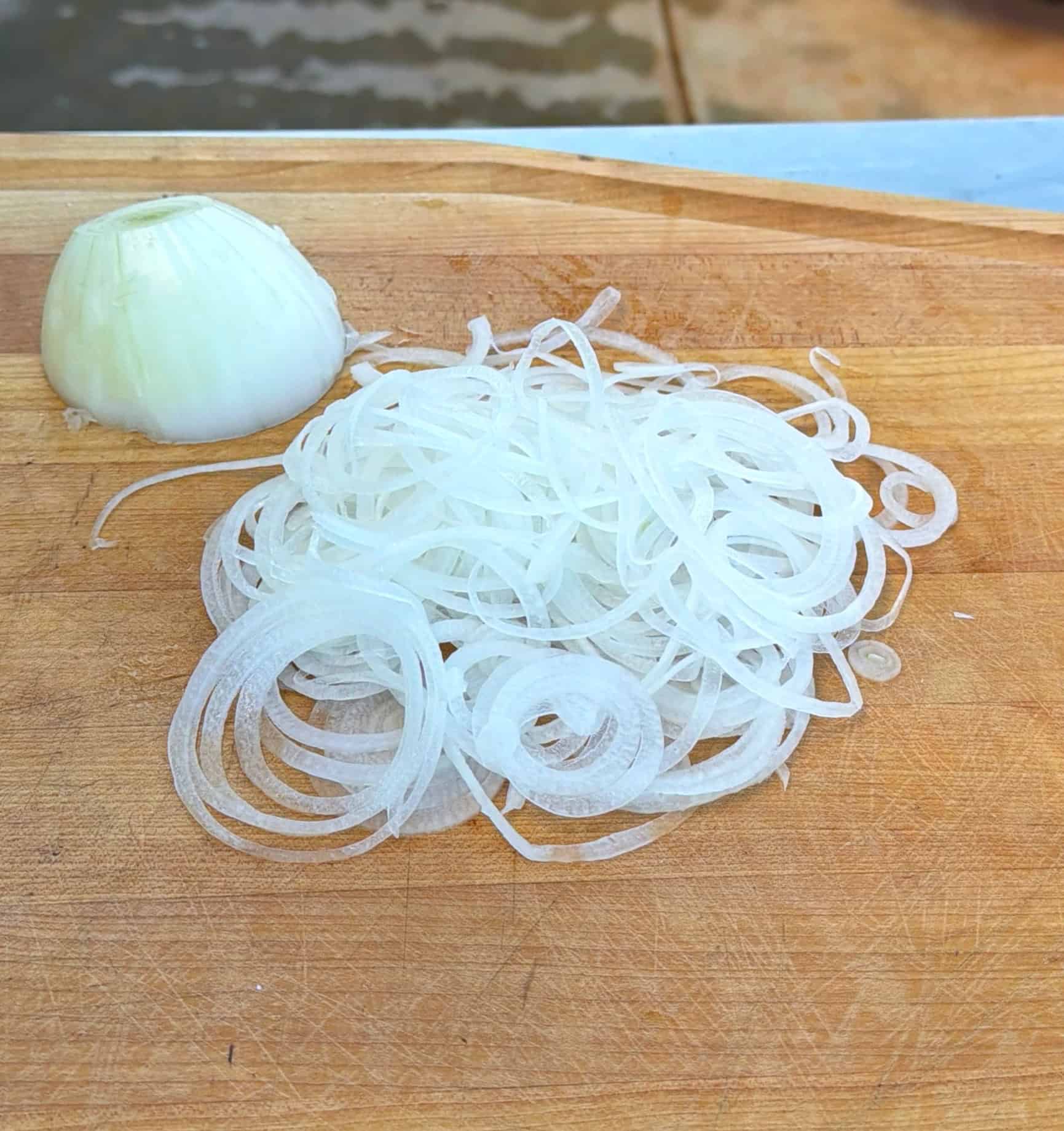 Thinly sliced white onion rings and a half onion rest on a wooden cutting board.