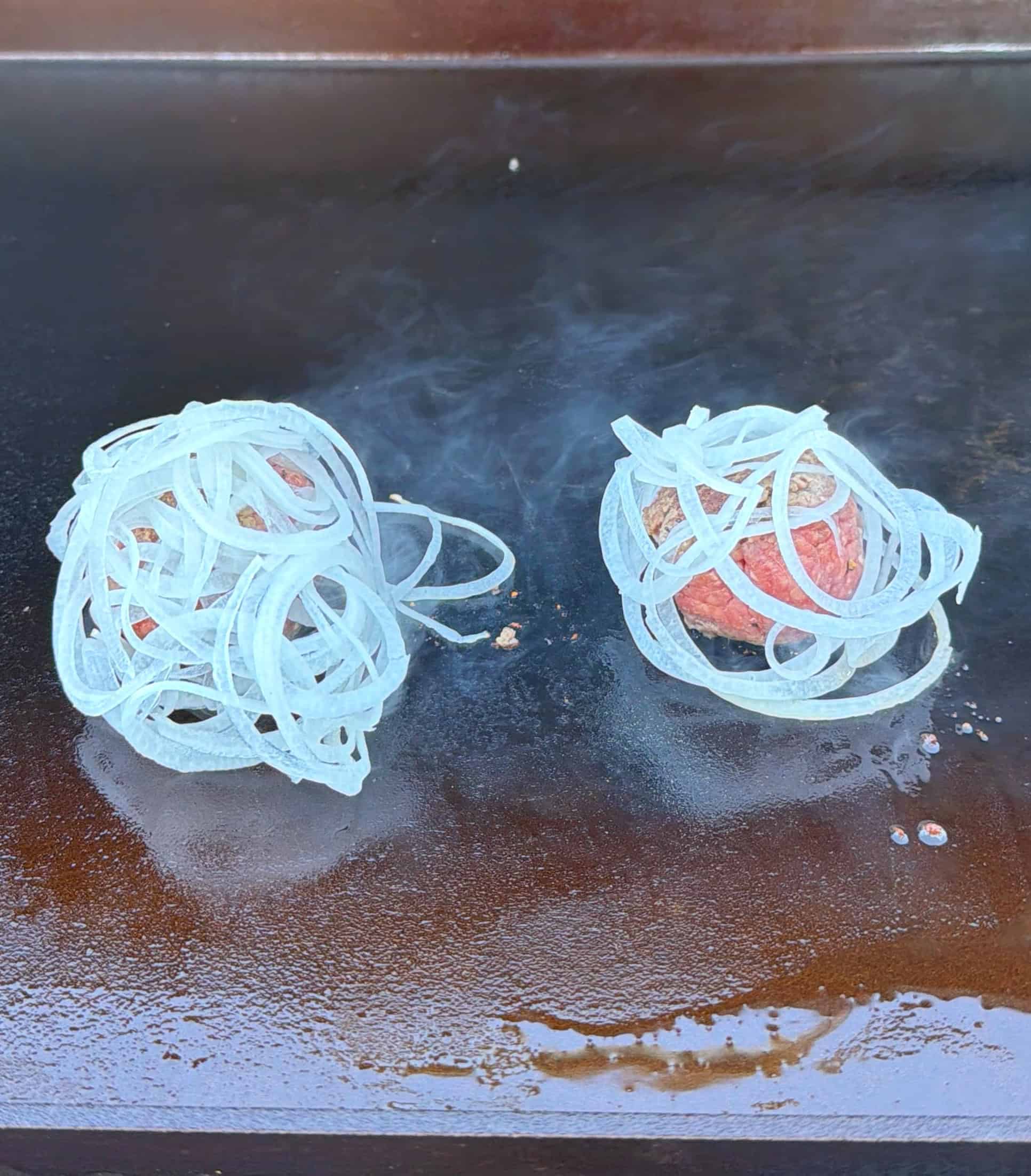 Two raw burger patties topped with piles of sliced onions are cooking on a flat top grill, with a bit of steam rising around them.