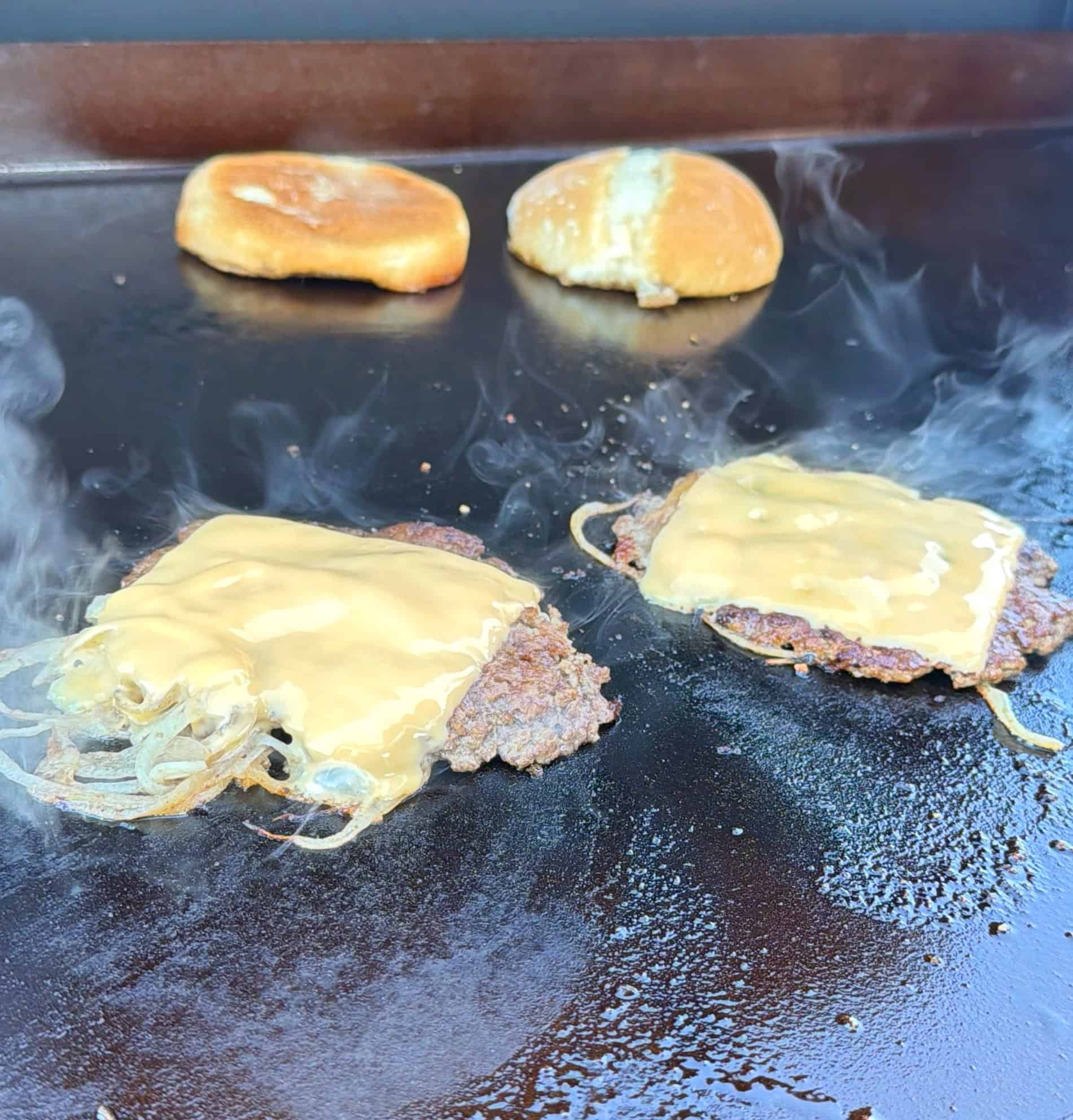 Two beef patties with melted cheese and grilled onions sizzle on a flat-top grill, with steam rising. Two burger buns are toasting in the background.