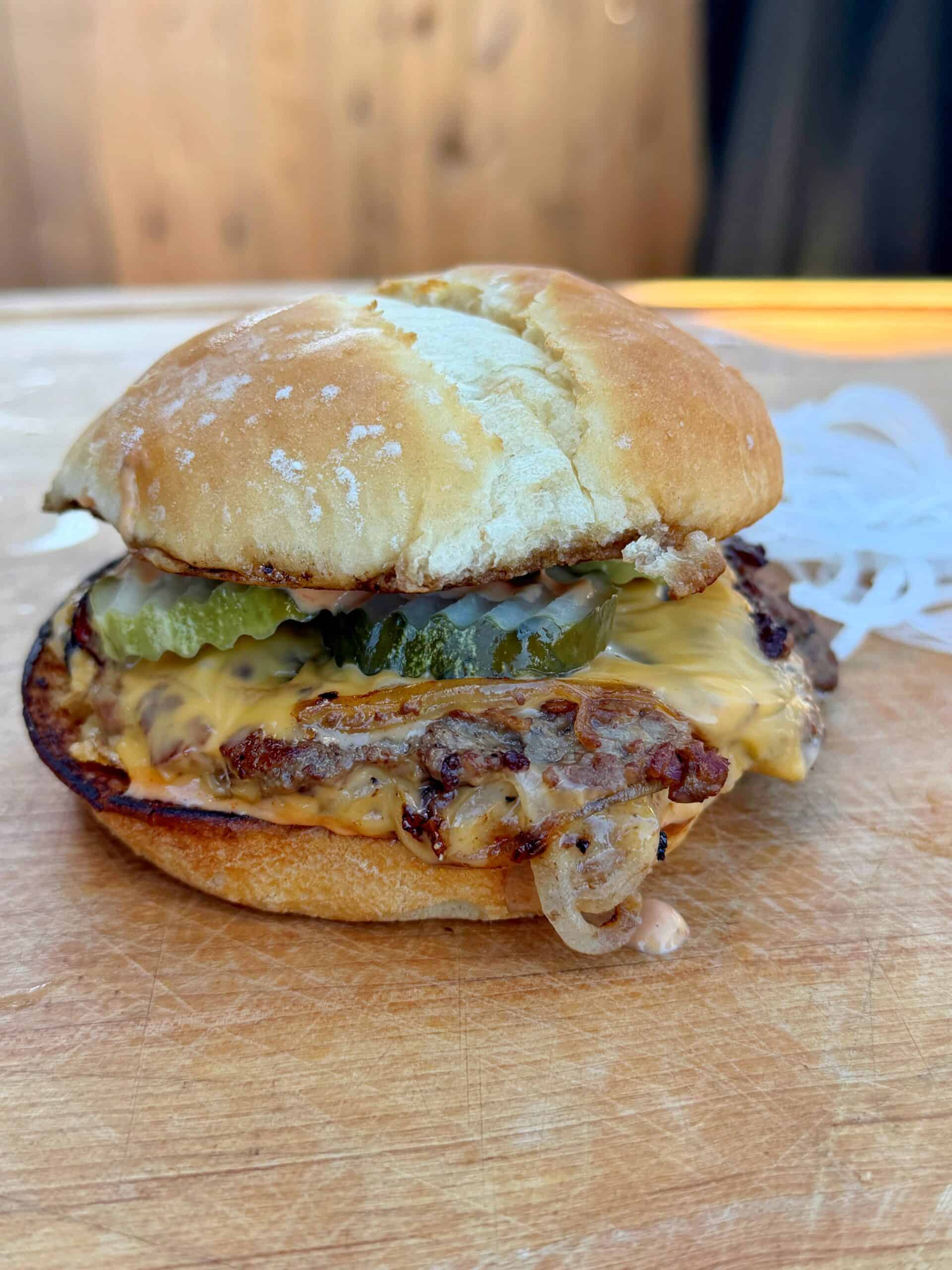 A cheeseburger on a wooden surface, topped with melted cheese, pickles, grilled onions, and served in a toasted bun. Thinly sliced raw onions are visible in the background.