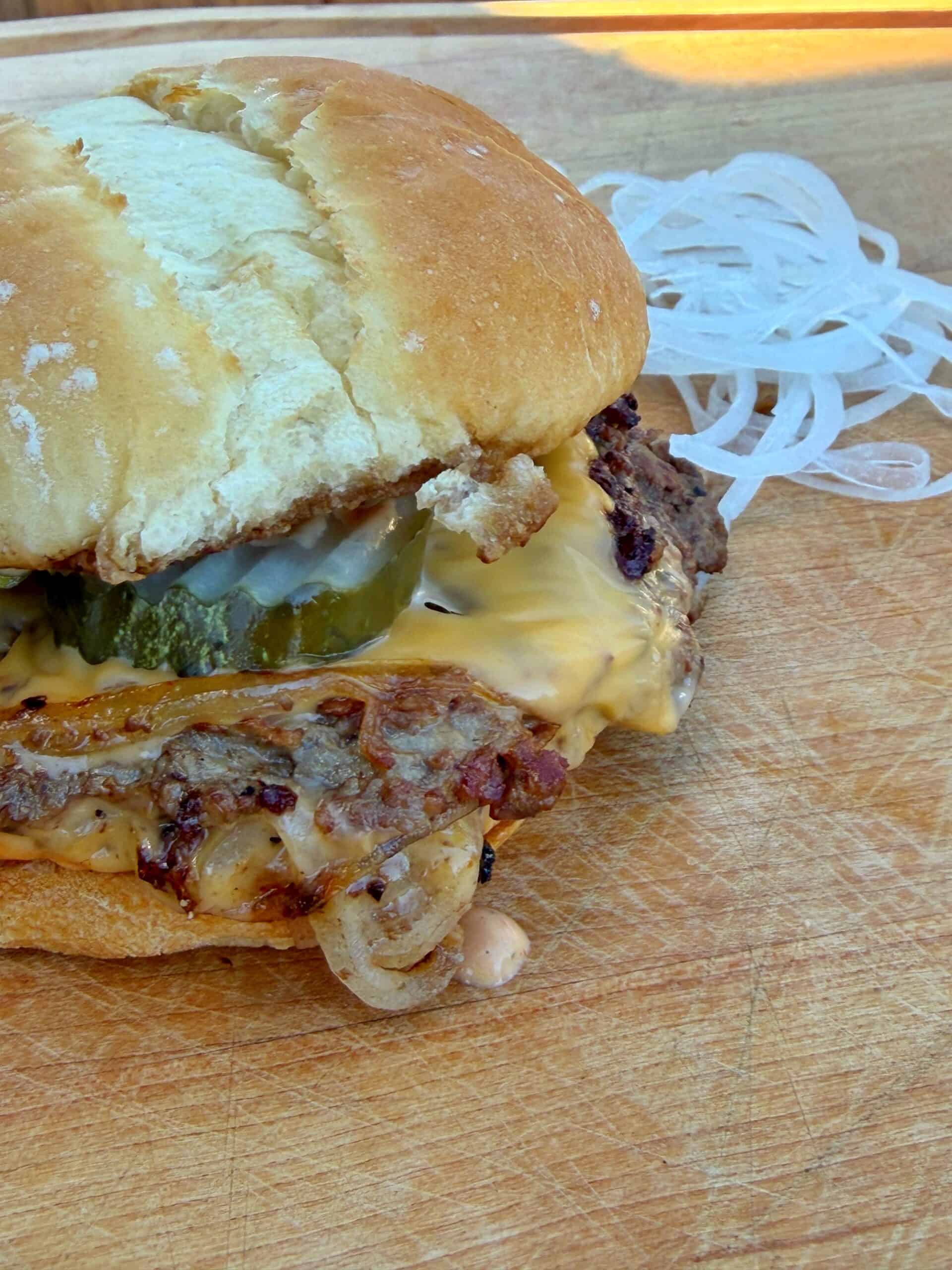 A close-up of an oklahoma smash burger cheeseburger with pickles, melted cheese, and grilled onions on a toasted bun, resting on a wooden surface. A small pile of raw, white onion slices is beside the sandwich.