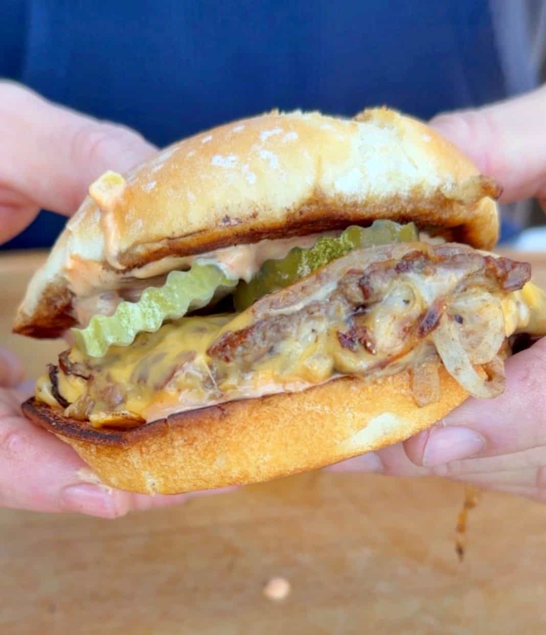 Close-up of hands holding a cheeseburger with melted cheese, grilled onions, pickles, and sauce inside a toasted bun.