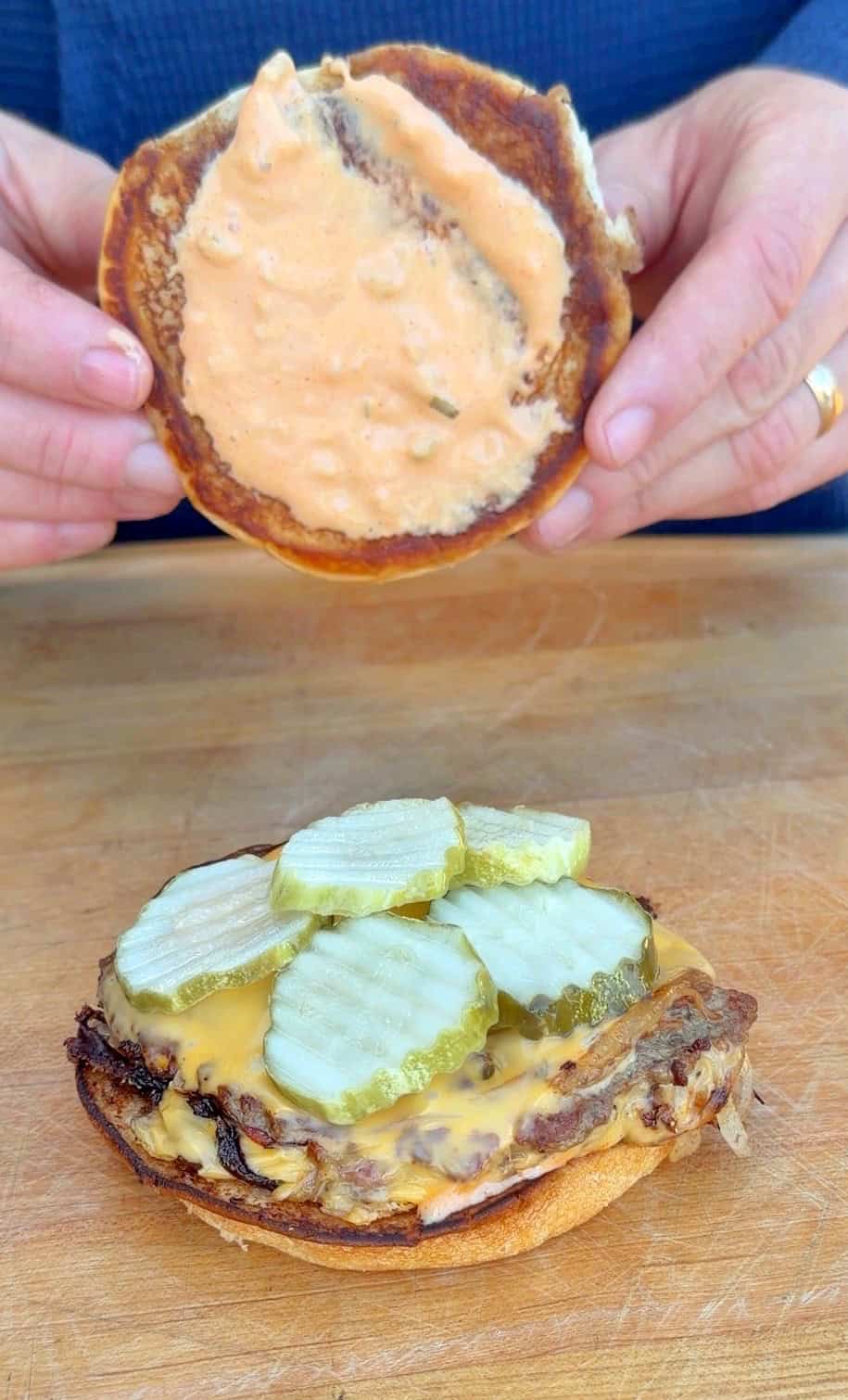 A person holds the top bun of a burger covered in a light orange sauce above the bottom bun, which has melted cheese, a beef patty, and sliced pickles, all placed on a wooden surface.