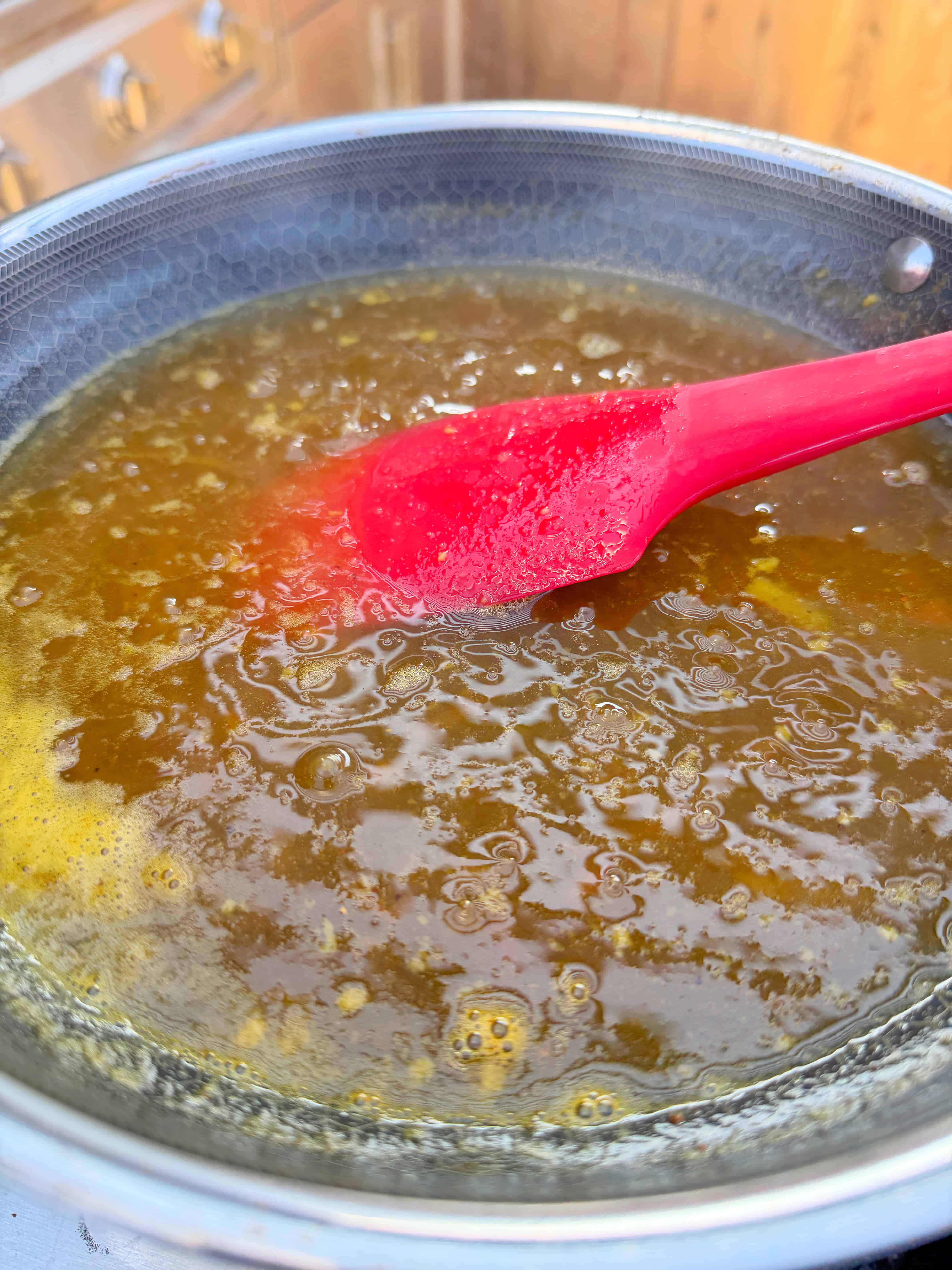 A close-up of a saucepan filled with bubbling golden liquid, being stirred with a red silicone spatula. The kitchen background is slightly blurred.