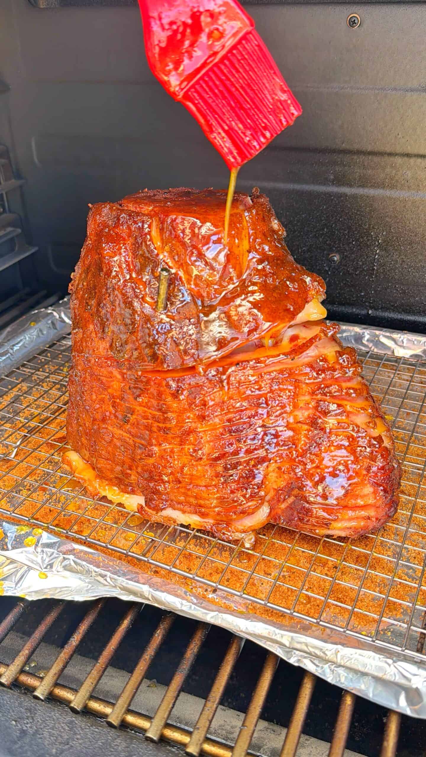 A large, glazed ham sits on a wire rack inside a smoker, covered in a shiny sauce. A red brush is being used to spread more glaze over the ham. Foil lines the smoker’s bottom for easy cleanup.