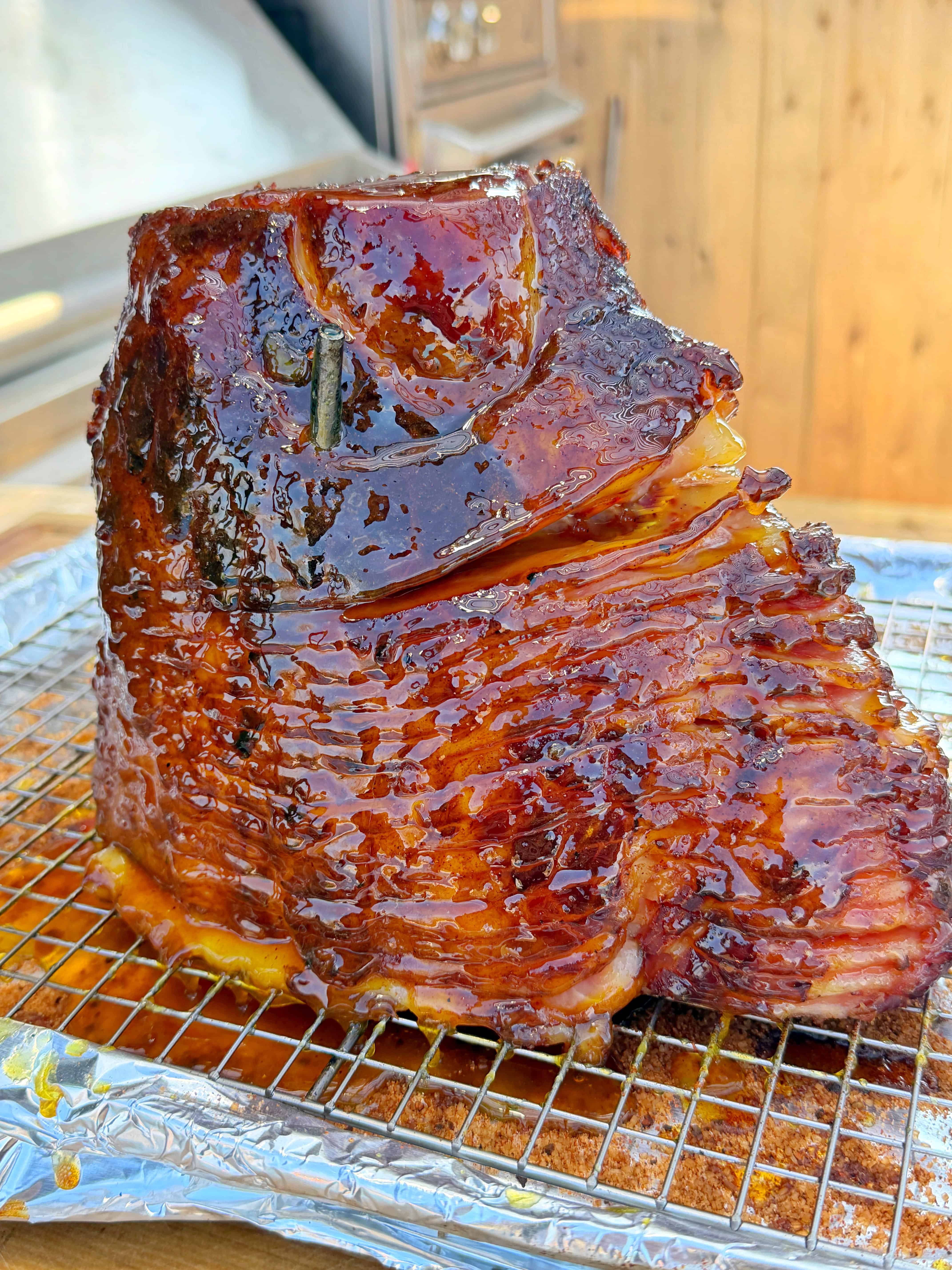 A glazed, spiral-cut ham sits on a wire rack covered with foil, glistening with caramelized glaze. The background shows blurred wooden panels, suggesting an outdoor setting.