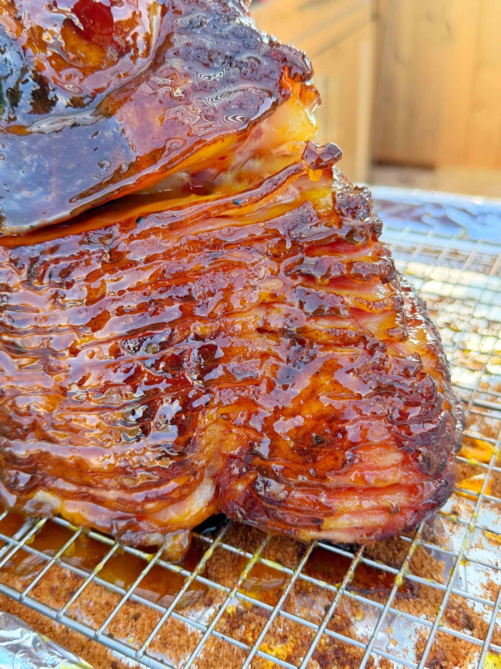 Close-up of a  orange glazed ham, spiral-cut ham resting on a wire rack, with a shiny caramelized surface and visible grill marks. The background is slightly blurred, focusing attention on the delicious, juicy meat.
