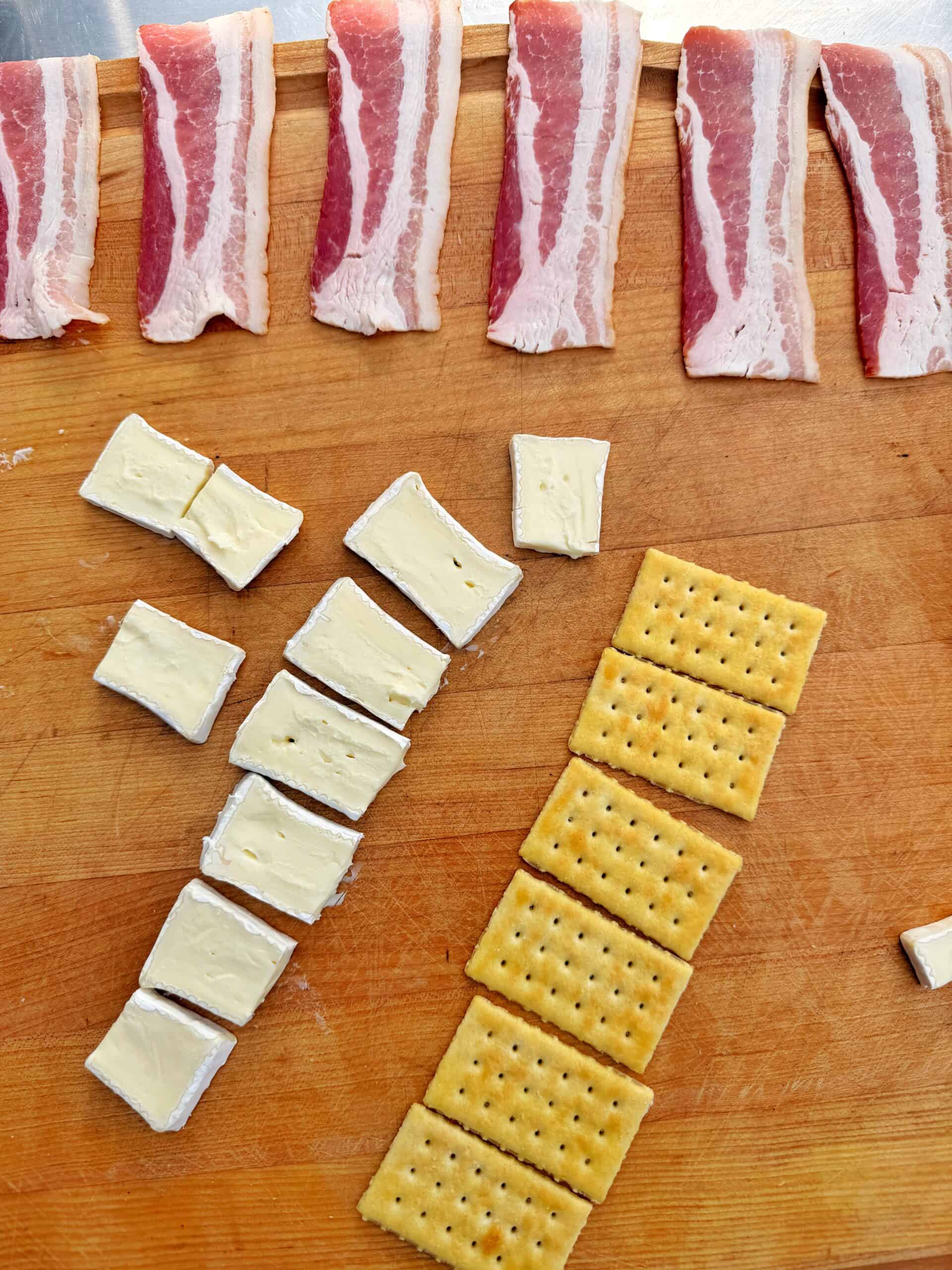 Slices of raw bacon, pieces of brie cheese, and rectangular crackers are neatly arranged on a wooden surface, with each ingredient in its own row. recipe is for bacon wrapped brie on crackers