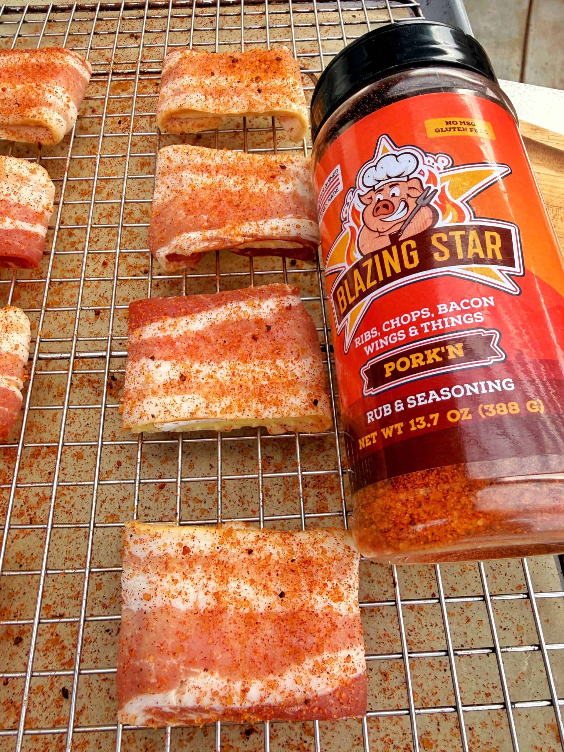 Rectangular pieces of raw pork belly, coated with red seasoning, are arranged on a wire rack. A container of Blazing Star PorkN Rub & Seasoning is placed on the right side of the rack.