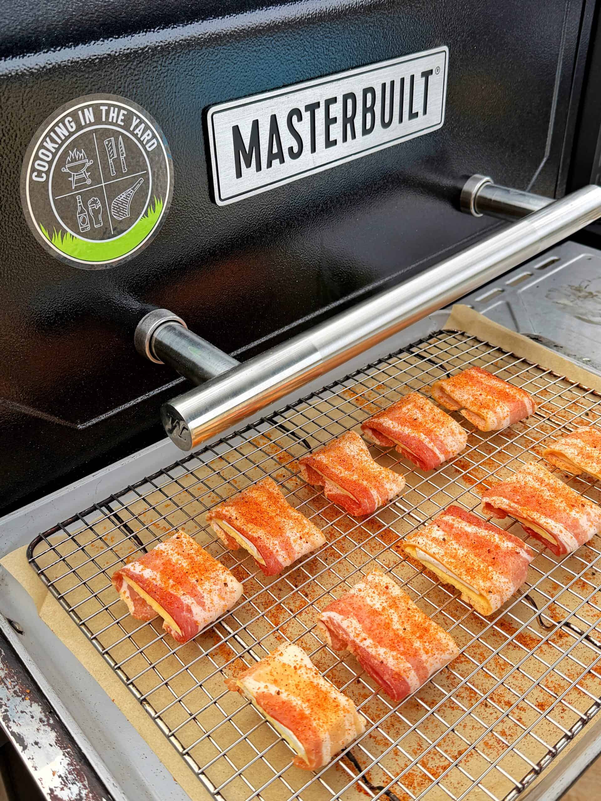 Several pieces of seasoned pork belly are arranged on a wire rack inside a Masterbuilt smoker. The rack rests on parchment paper, and the smoker lid is partially open, showing a Cooking in the Yard sticker.