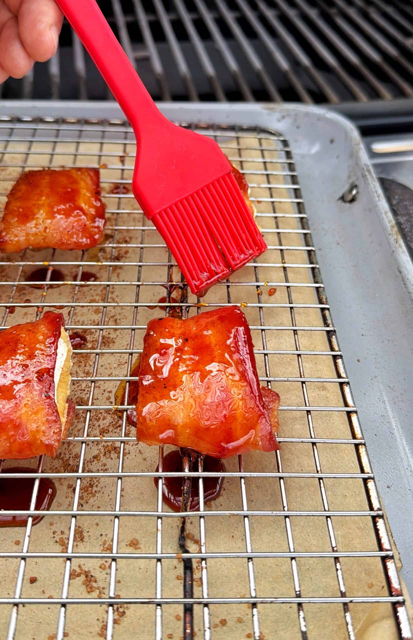 A hand uses a red brush to apply glaze to pieces of bacon-wrapped food on a wire rack over a baking sheet, set on an outdoor grill.