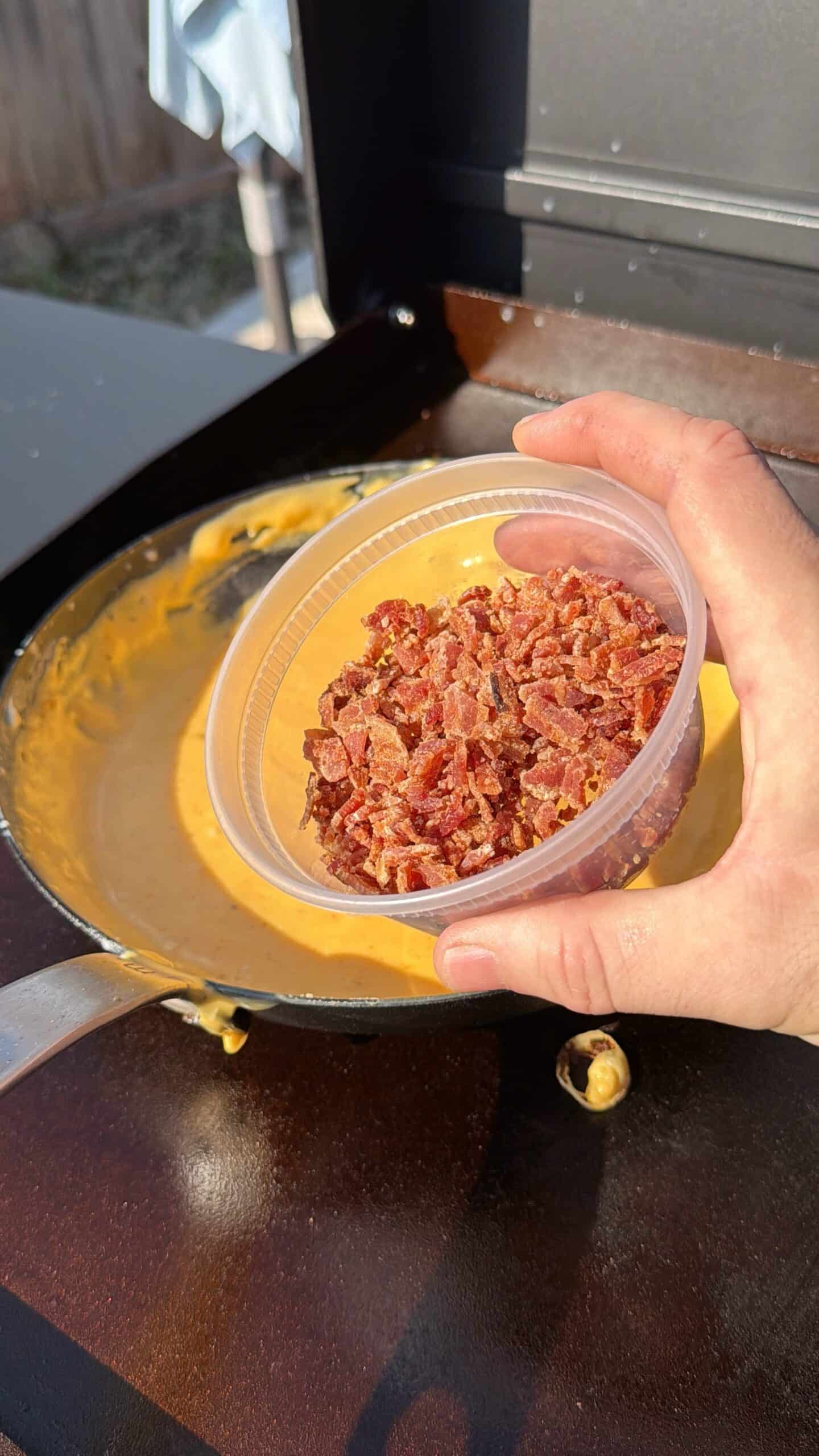 A hand holds a plastic container of chopped bacon over a pan of melted cheese sauce on an outdoor grill, ready to add the bacon to the cheese mixture.