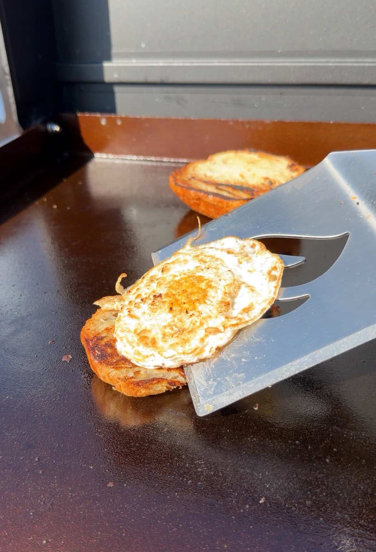 A metal spatula lifts a cooked egg on top of a slice of browned bread from a flat griddle, with another piece of bread in the background.
