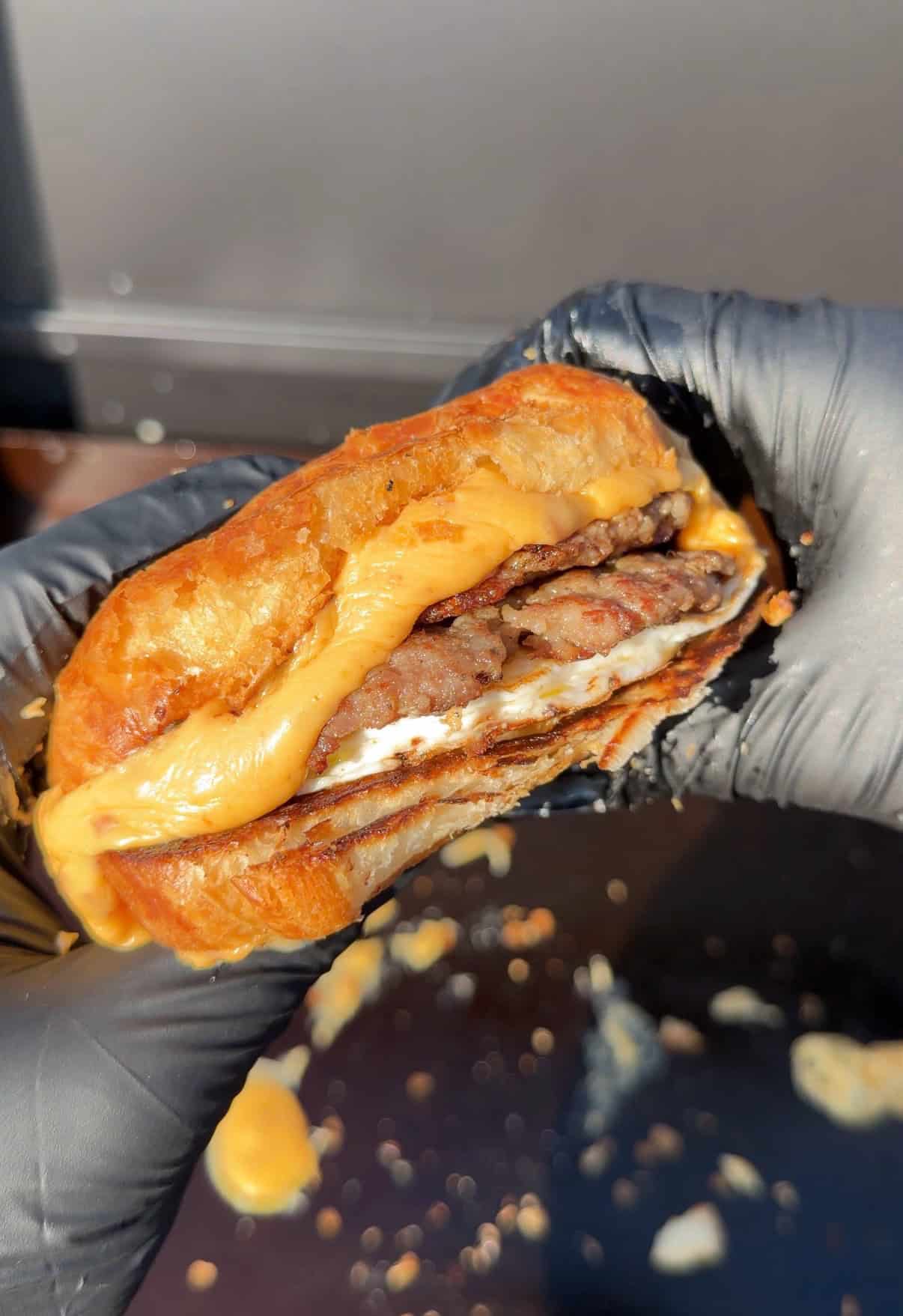 A close-up of a gloved hand holding a bacon queso breakfast sandwich with melted cheese, grilled beef patties, and a crispy, golden bun, with cheese oozing out and crumbs scattered below.
