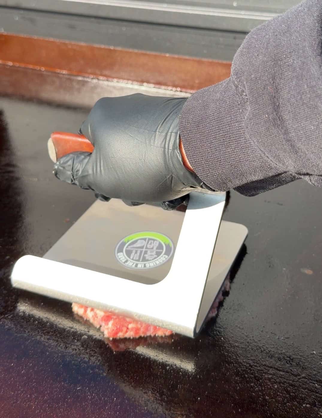 A person wearing a black glove presses down on a spatula, smashing a raw burger patty on a flat-top grill.