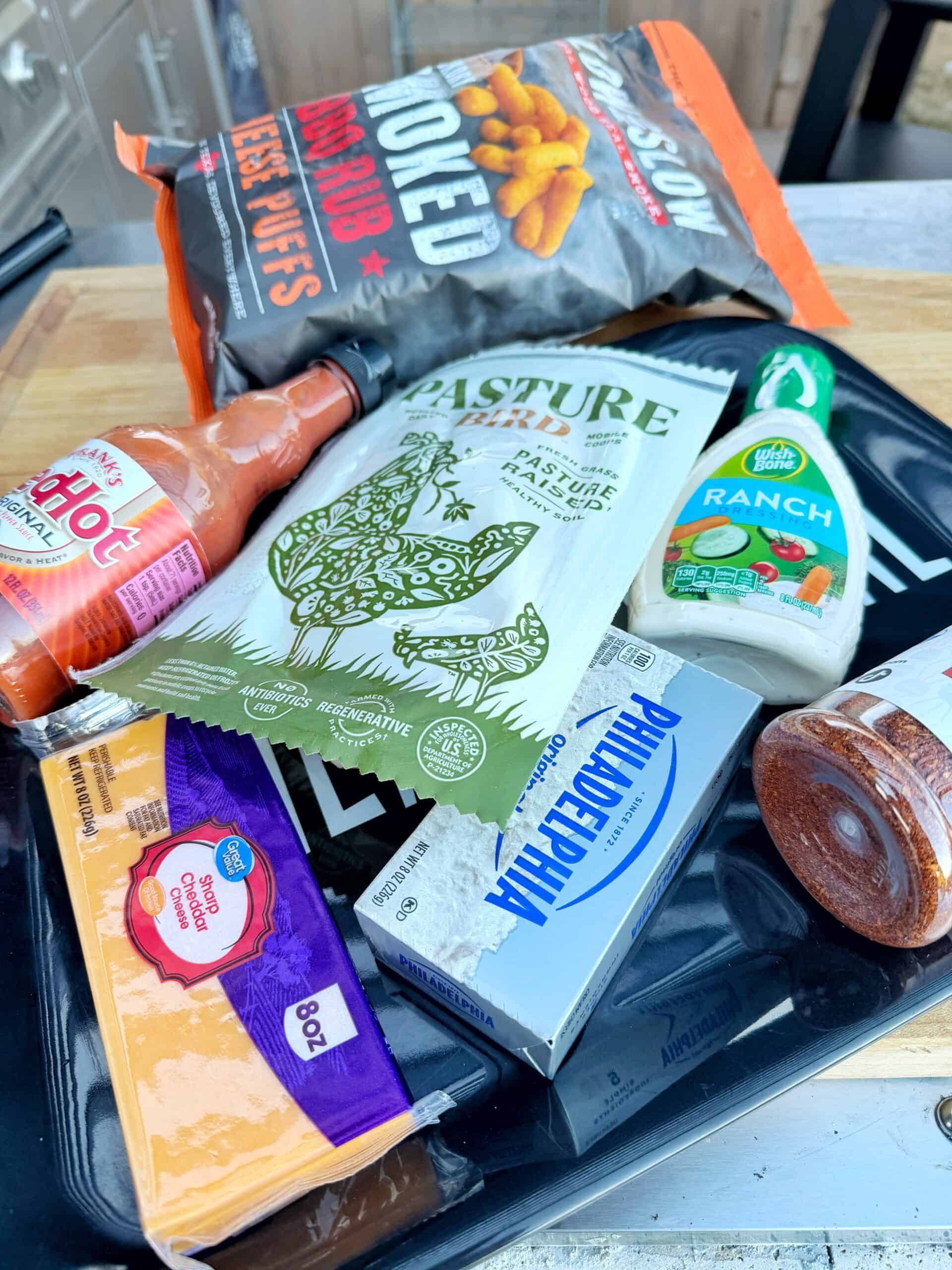 Ingredients for buffalo chicken dip: A variety of foods on a cutting board, including Frank’s RedHot sauce, BBQ pork puffs, ranch dressing, shredded cheddar cheese, pasture-raised chicken breast, Philadelphia cream cheese, and a sausage.