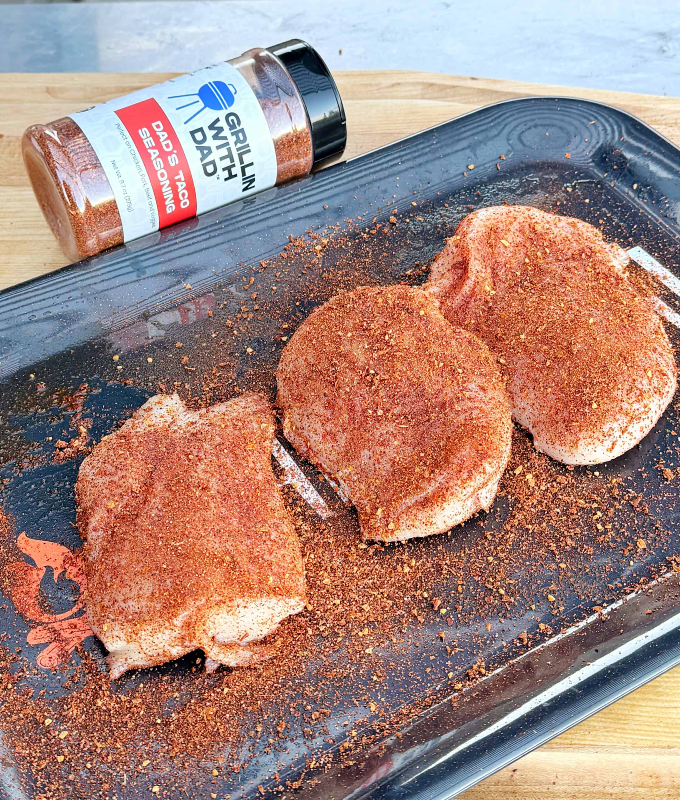 Three raw pork chops coated with BBQ seasoning are arranged on a black tray. A seasoning bottle labeled “Grillin’ With Dad BBQ Pro Seasoning” lies nearby on a wooden surface.