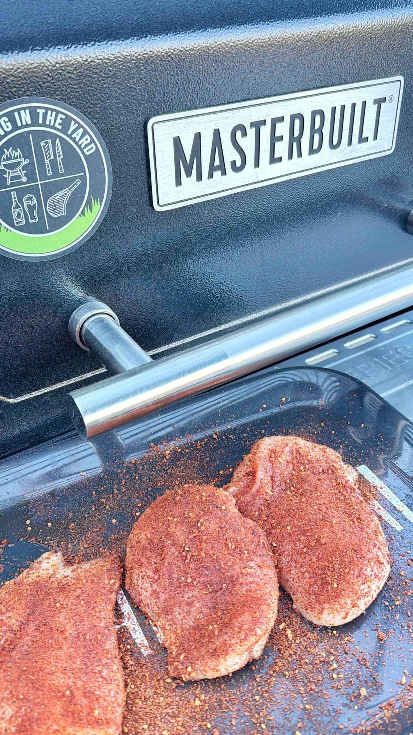 Three pieces of seasoned meat are placed on a grill surface under a MASTERBUILT branded smoker or grill lid, with seasoning spilled on the side. A circular sticker about yard grilling is visible in the background.