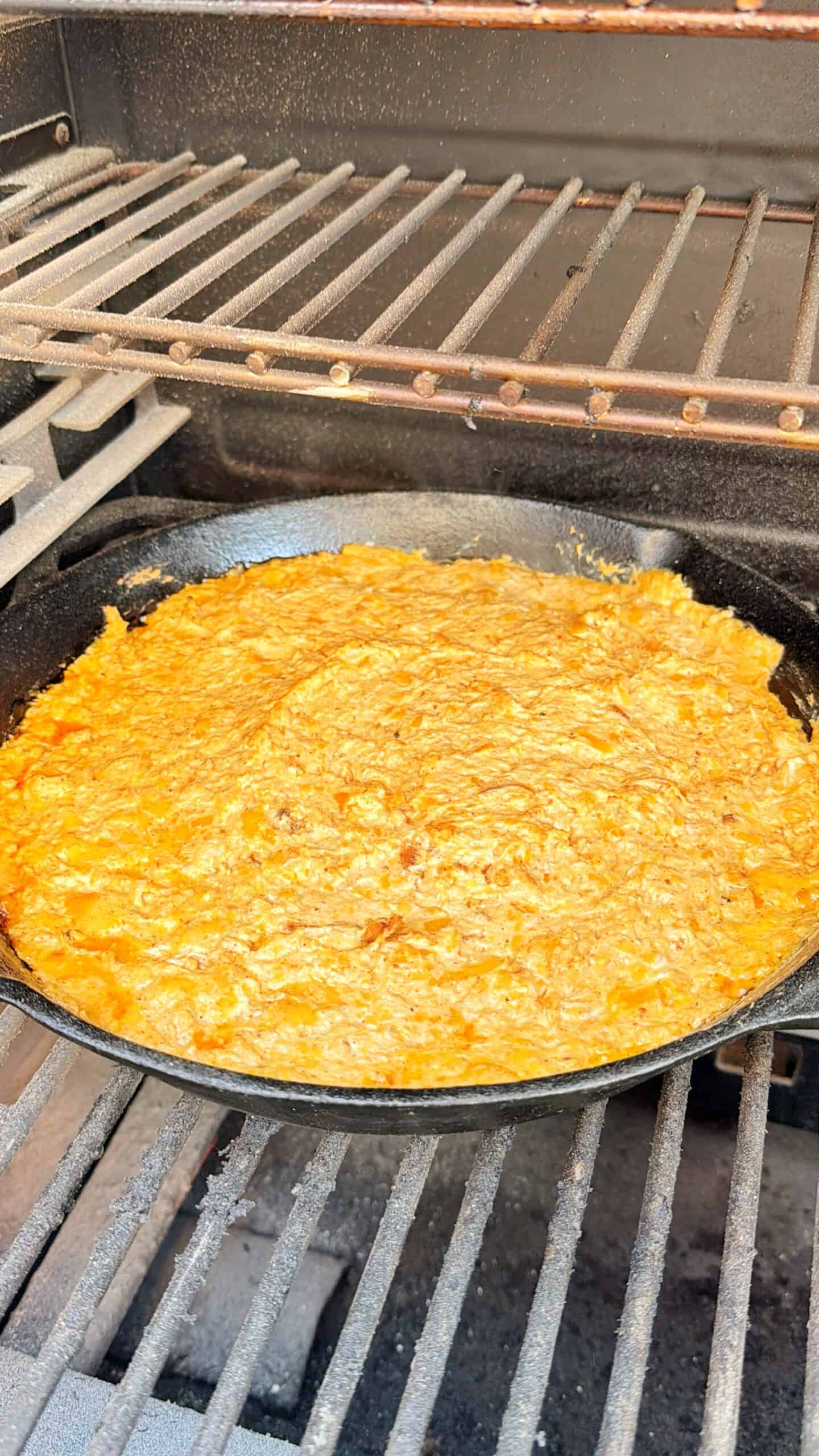 A cast iron skillet filled with a cheesy, orange-colored dip sits on the grill grates of an outdoor barbecue, cooking over the heat.