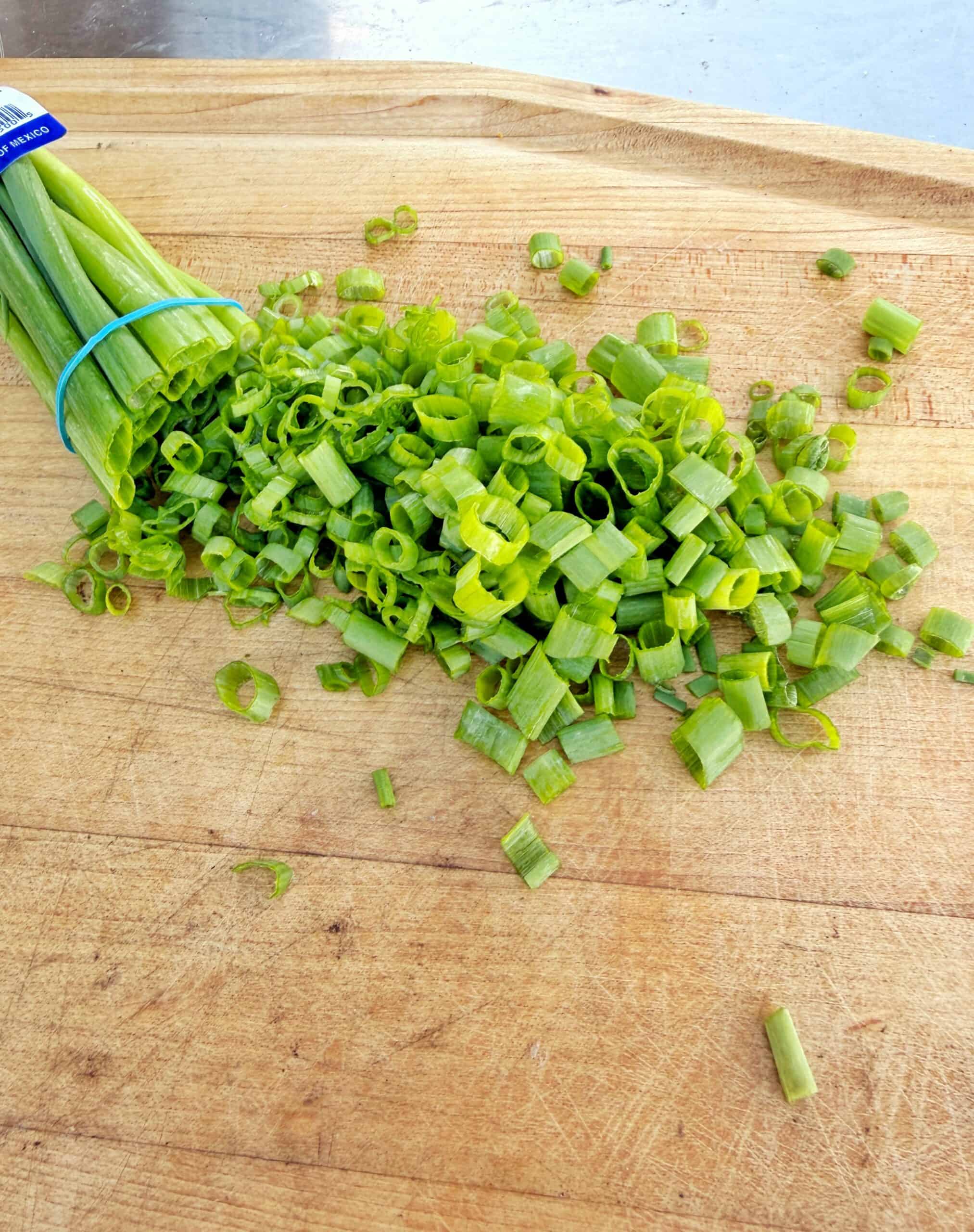 A bundle of fresh green onions lies on a wooden cutting board, with several slices of chopped green onion scattered around.